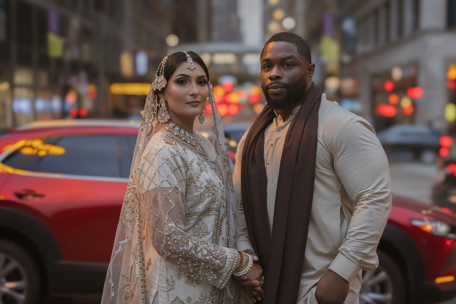 Interracial wedding couple in South Asian bridal attire pose holding hands on a vibrant city street.