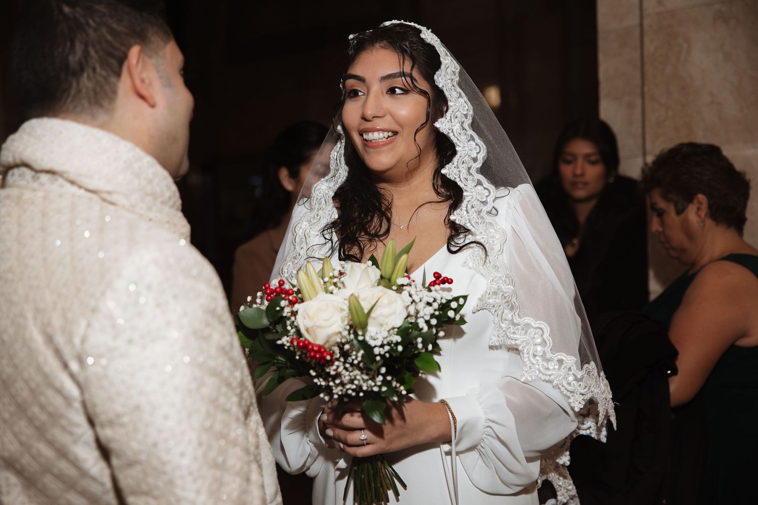 Smiling bride in white gown and lace veil holding red and white bouquet at wedding ceremony.