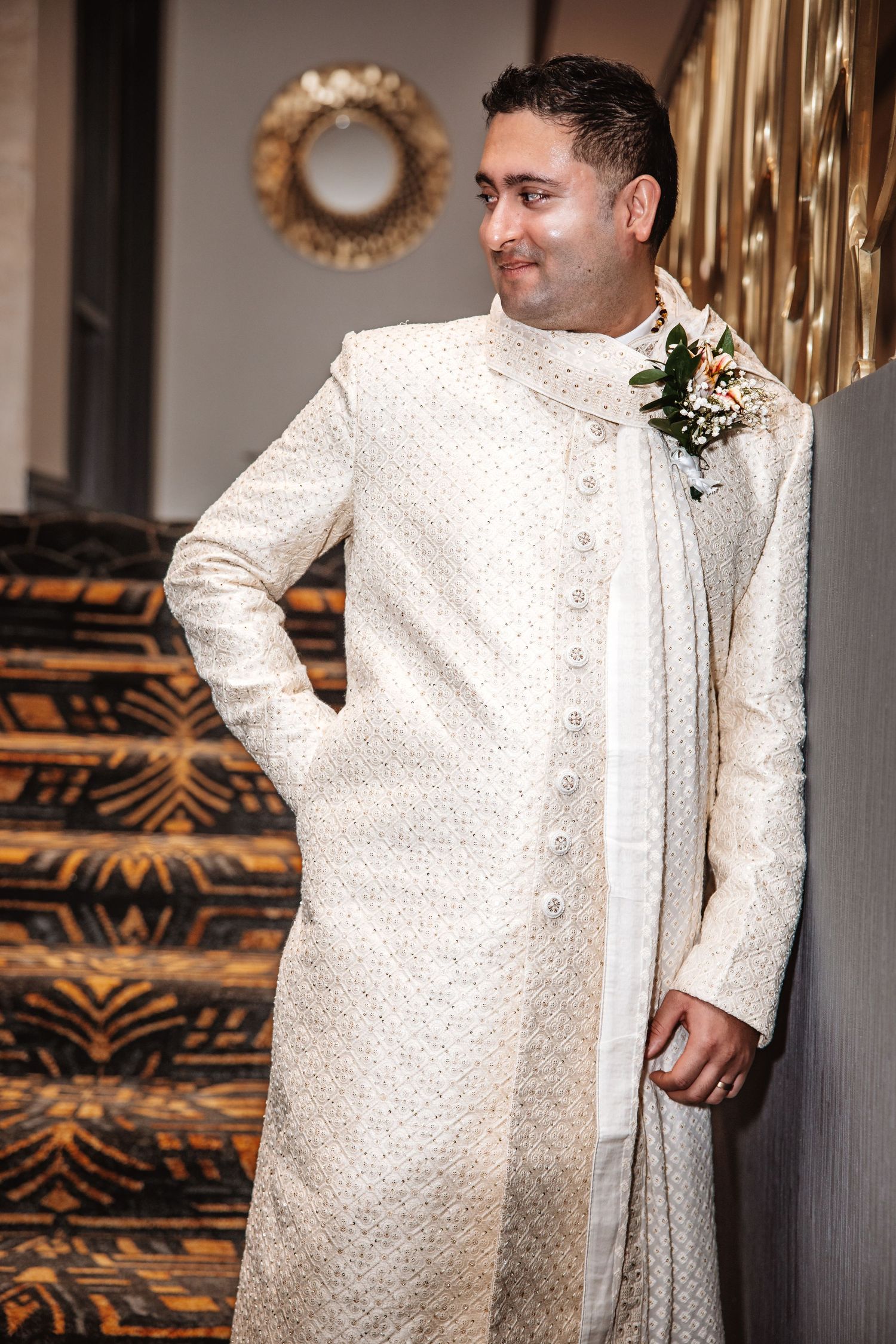 Elegant groom in ivory embroidered sherwani with boutonniere leans against wall by staircase.