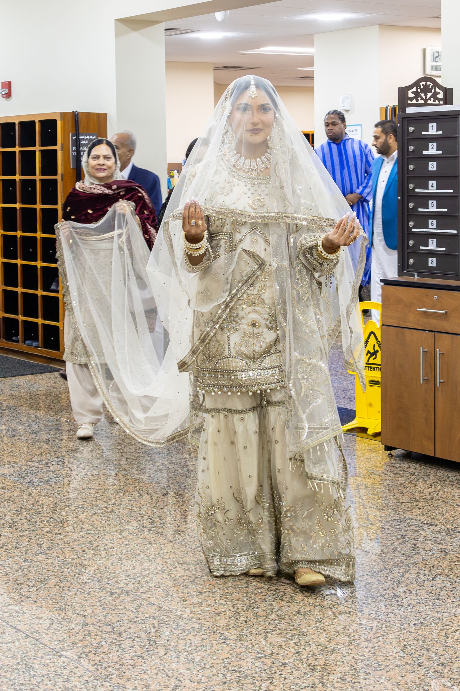 A South Asian bride in white embroidered lehenga walks through a mosque lobby holding her veil.