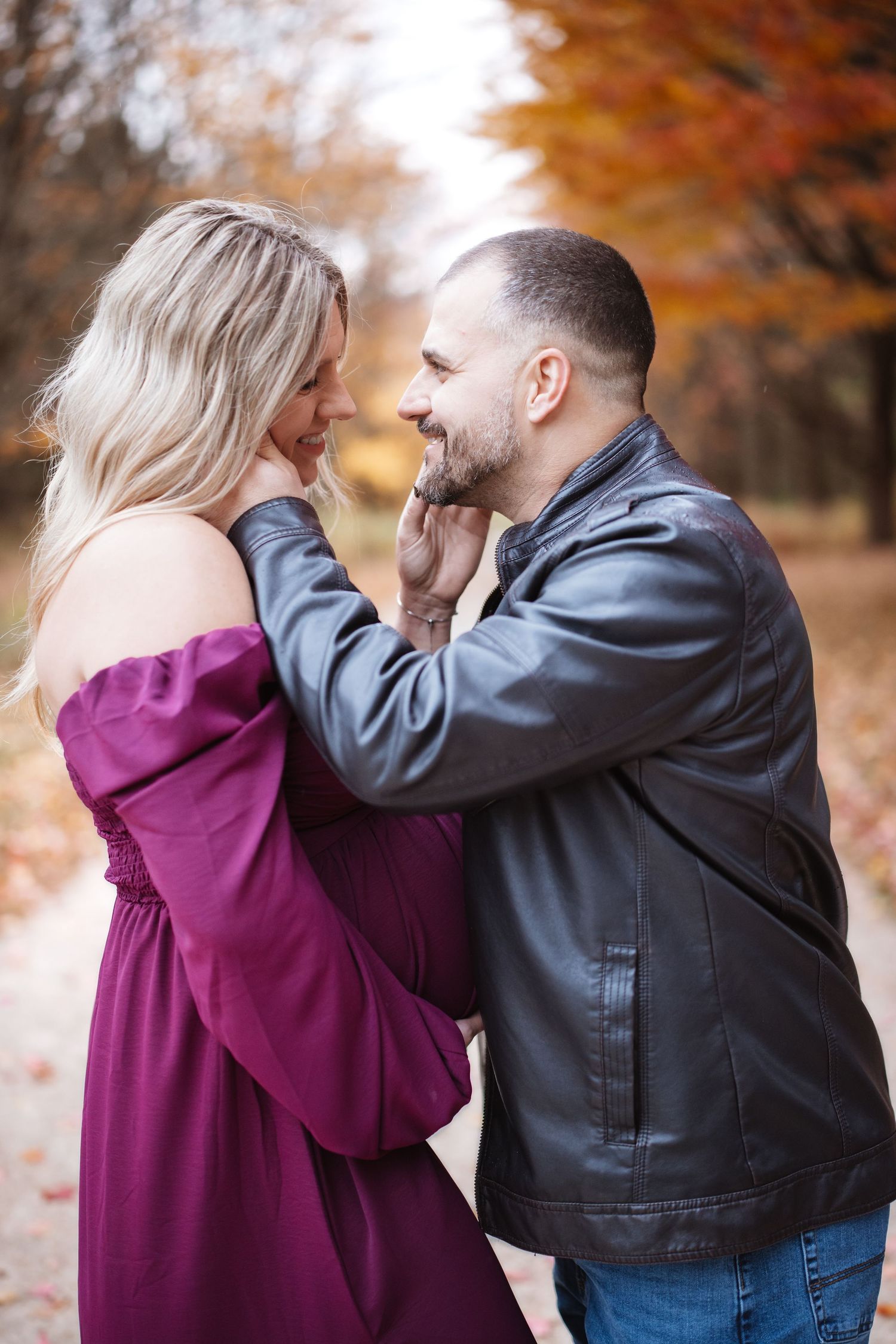 Laughing expectant couple face to face outdoors, man gently cups womans chin wearing leather jacket among fall foliage.