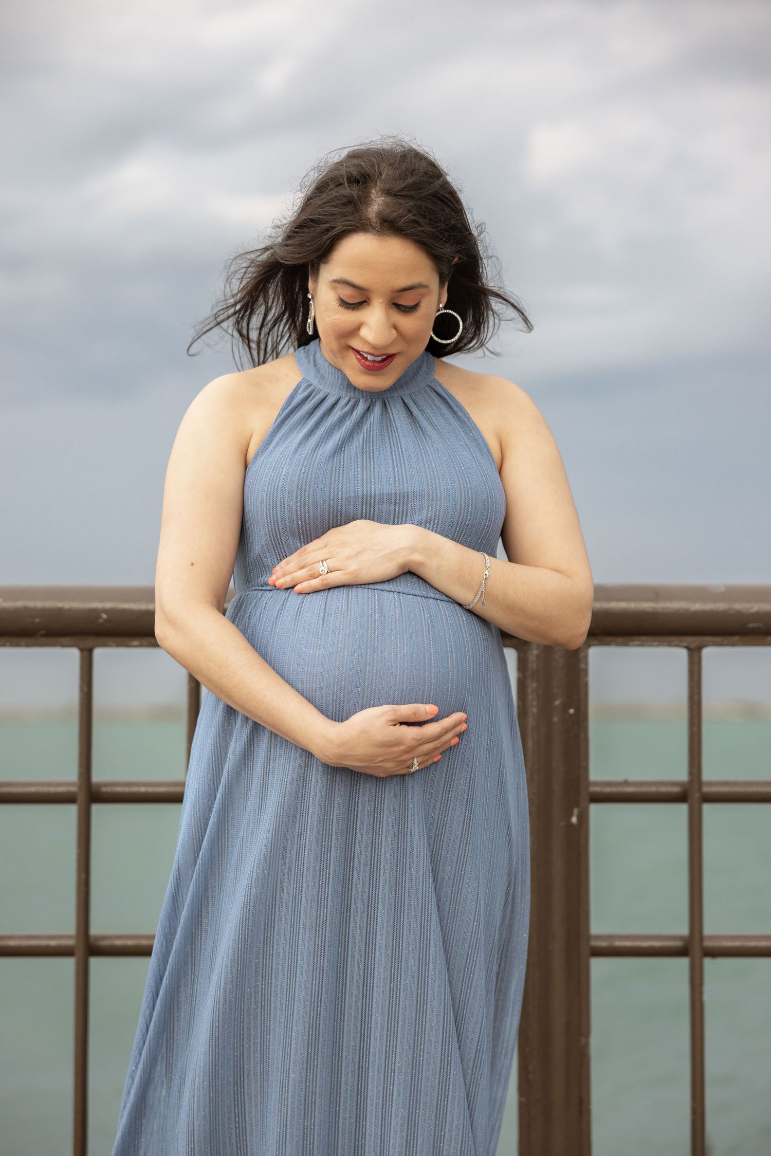 Joyful pregnant woman in blue pleated halter dress smiling down at her baby bump on a breezy pier.