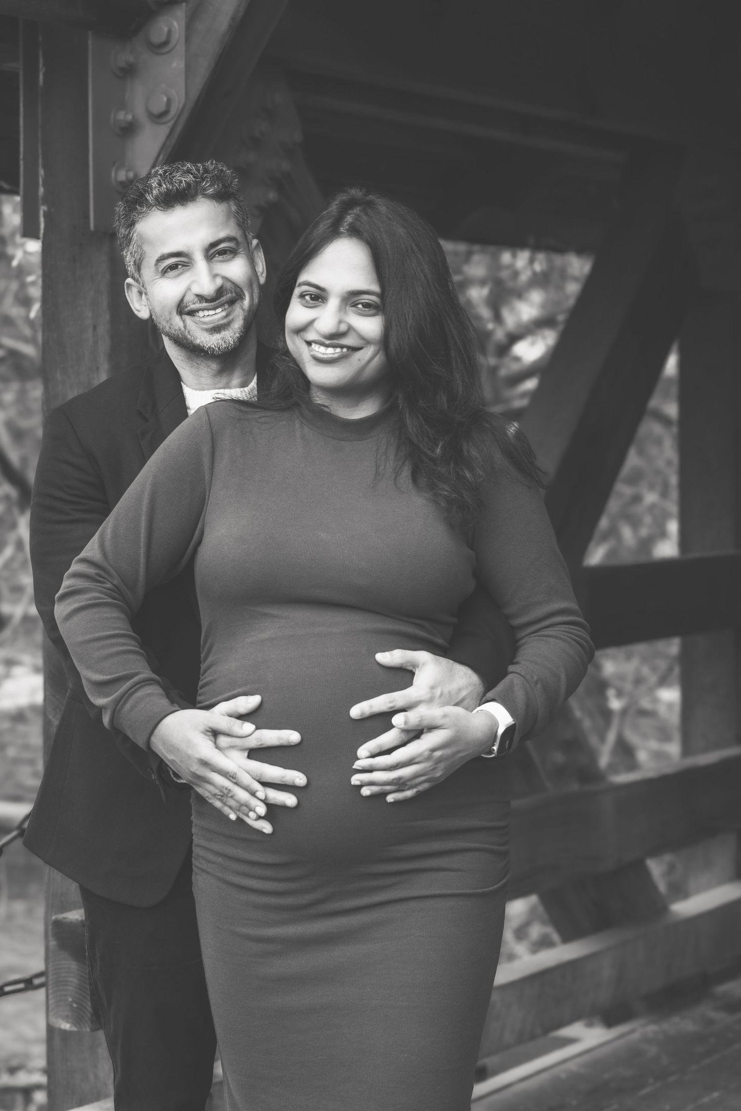 Black and white maternity photo of couple embracing under wooden bridge structure.
