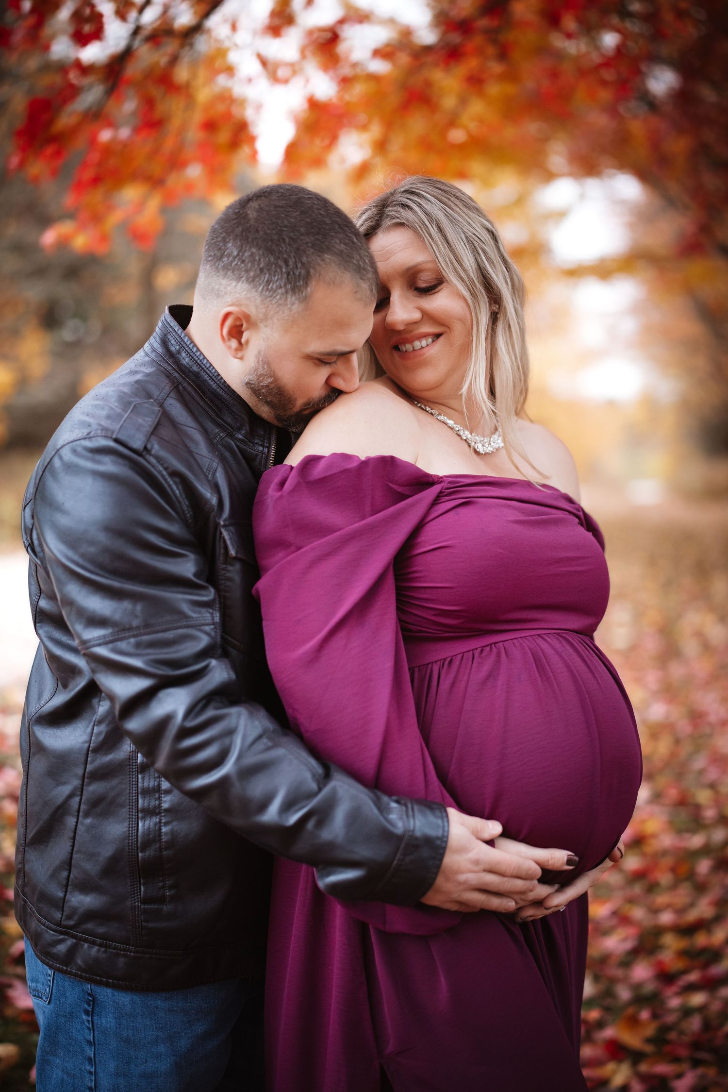 Couple embracing in colorful autumn park, pregnant woman in purple gown smiling with partner.