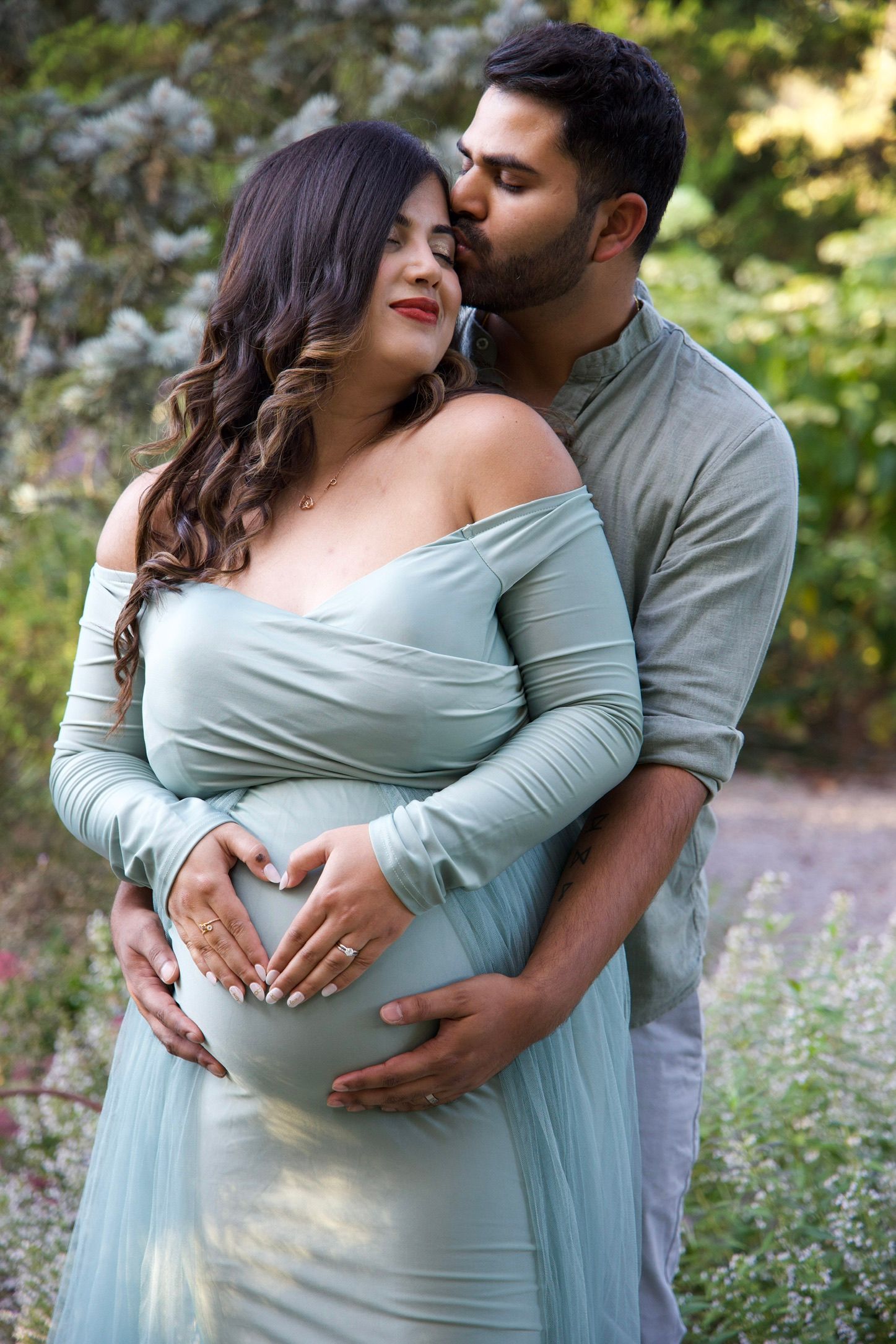 Couple sharing intimate maternity photo, man kissing womans head while forming heart shape on baby bump.