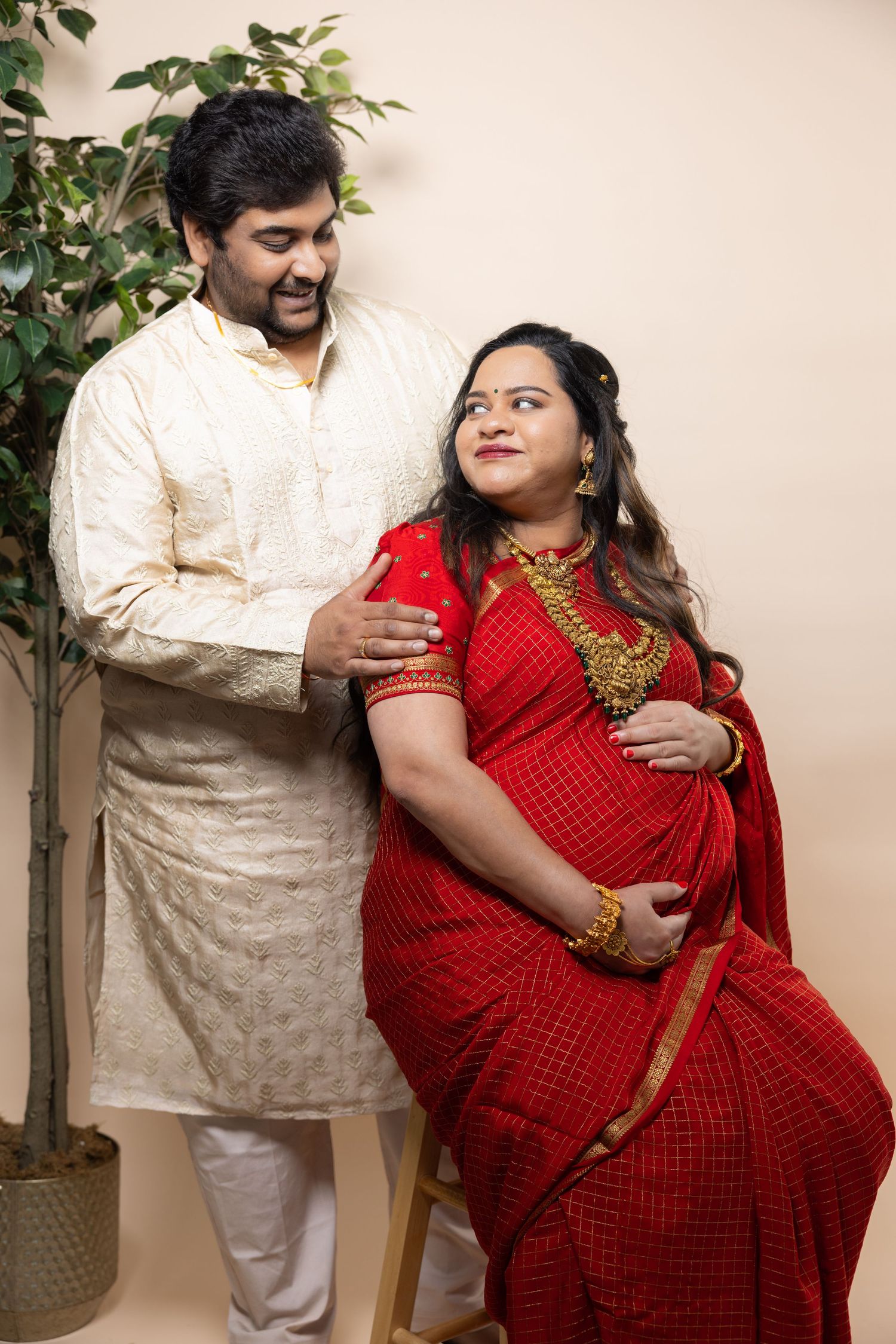 Indian couple in traditional attire, man in kurta with pregnant woman in red saree and gold jewelry.