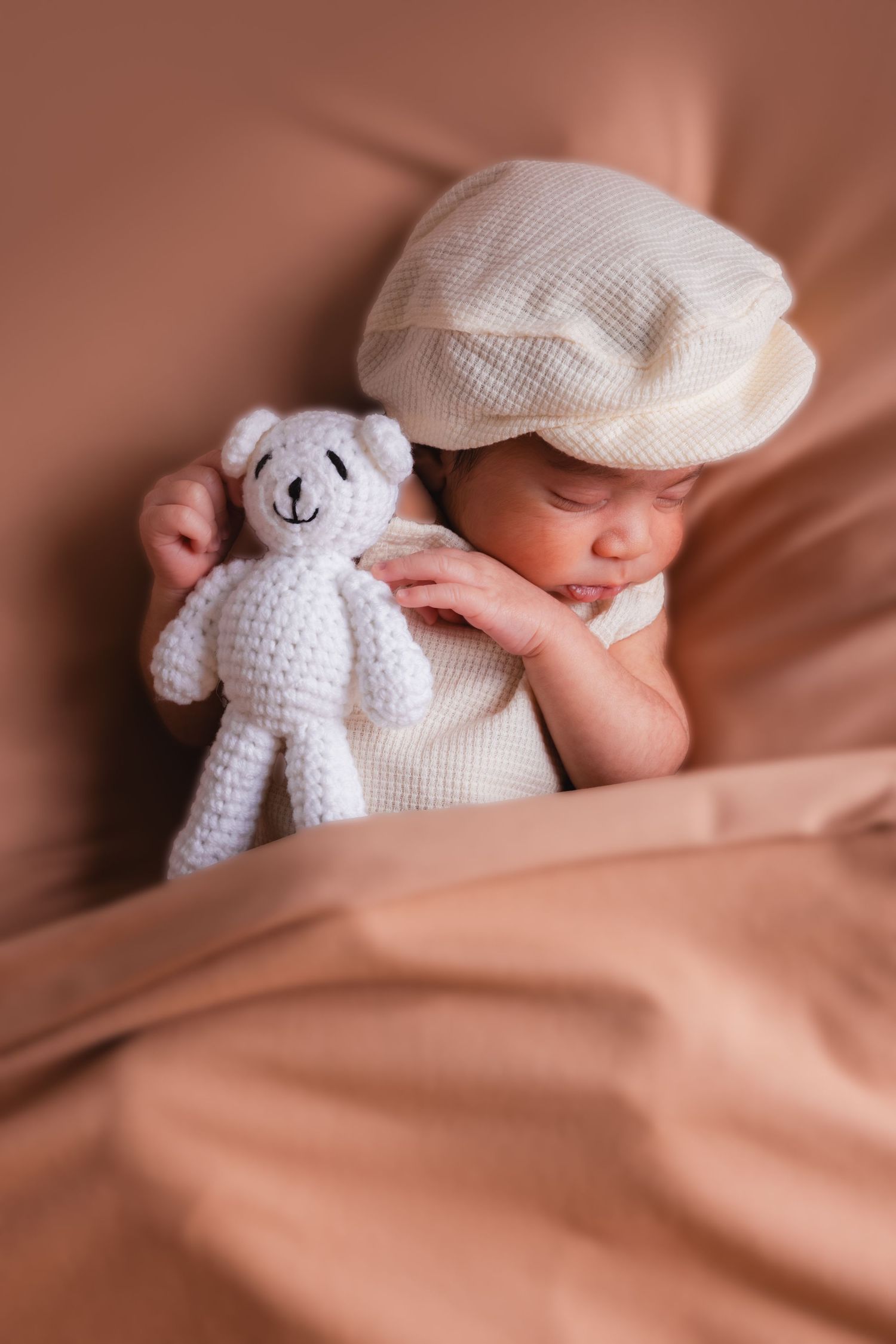 Sleeping newborn baby wearing white cap cuddles knitted teddy bear on beige blanket.