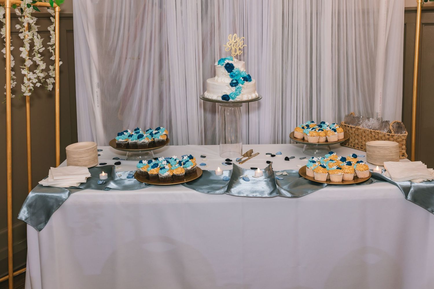Elegant wedding dessert table with tiered cake and blue-decorated cupcakes on silver cloth.