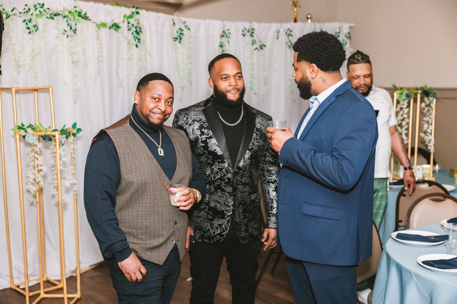Three well-dressed men in suits chat and laugh together at a decorated wedding reception venue.