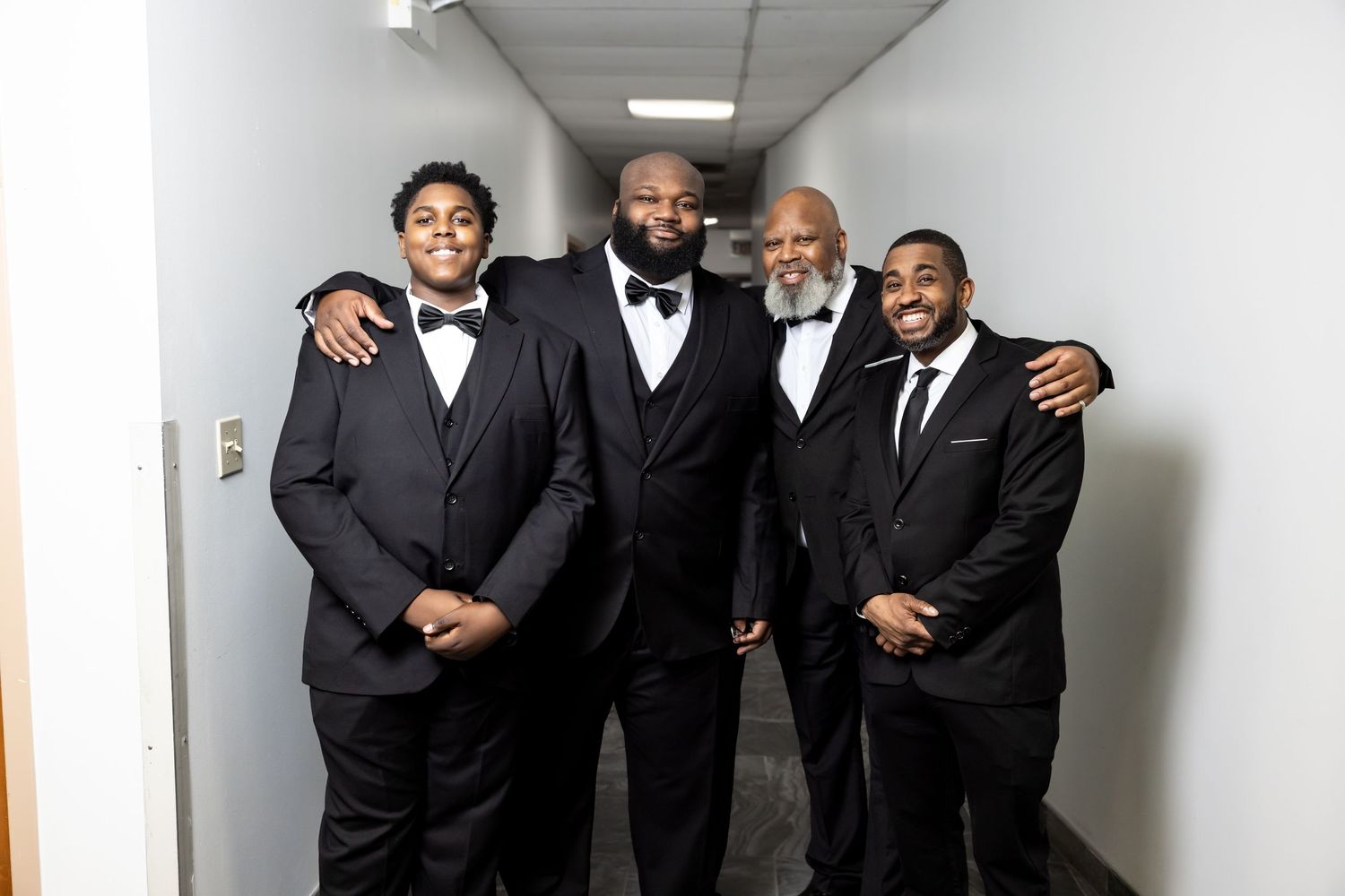 Four men in black tuxedos smiling and posing together in a hallway at a wedding.