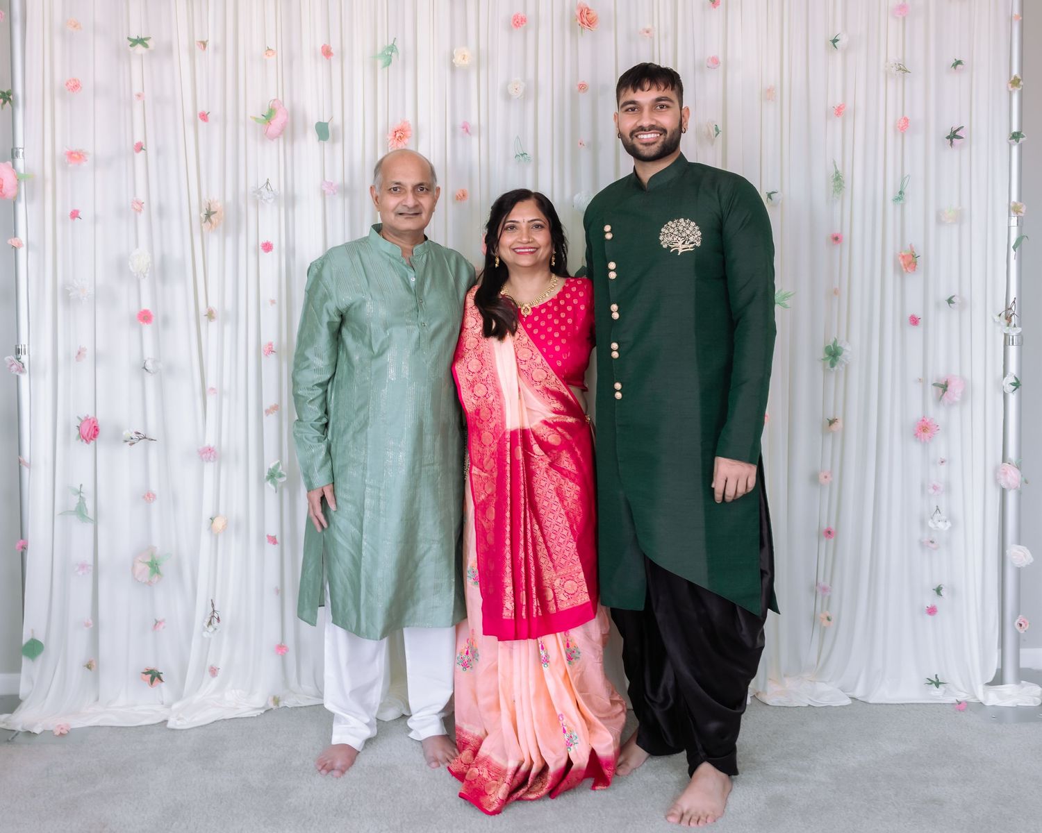 Three family members in traditional Indian attire pose together at festive event with floral backdrop.