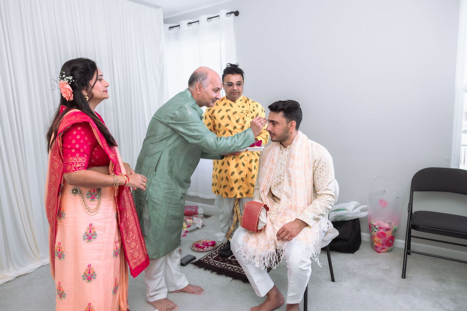 Elder man blessing seated groom during Indian wedding ceremony while woman in pink saree watches.