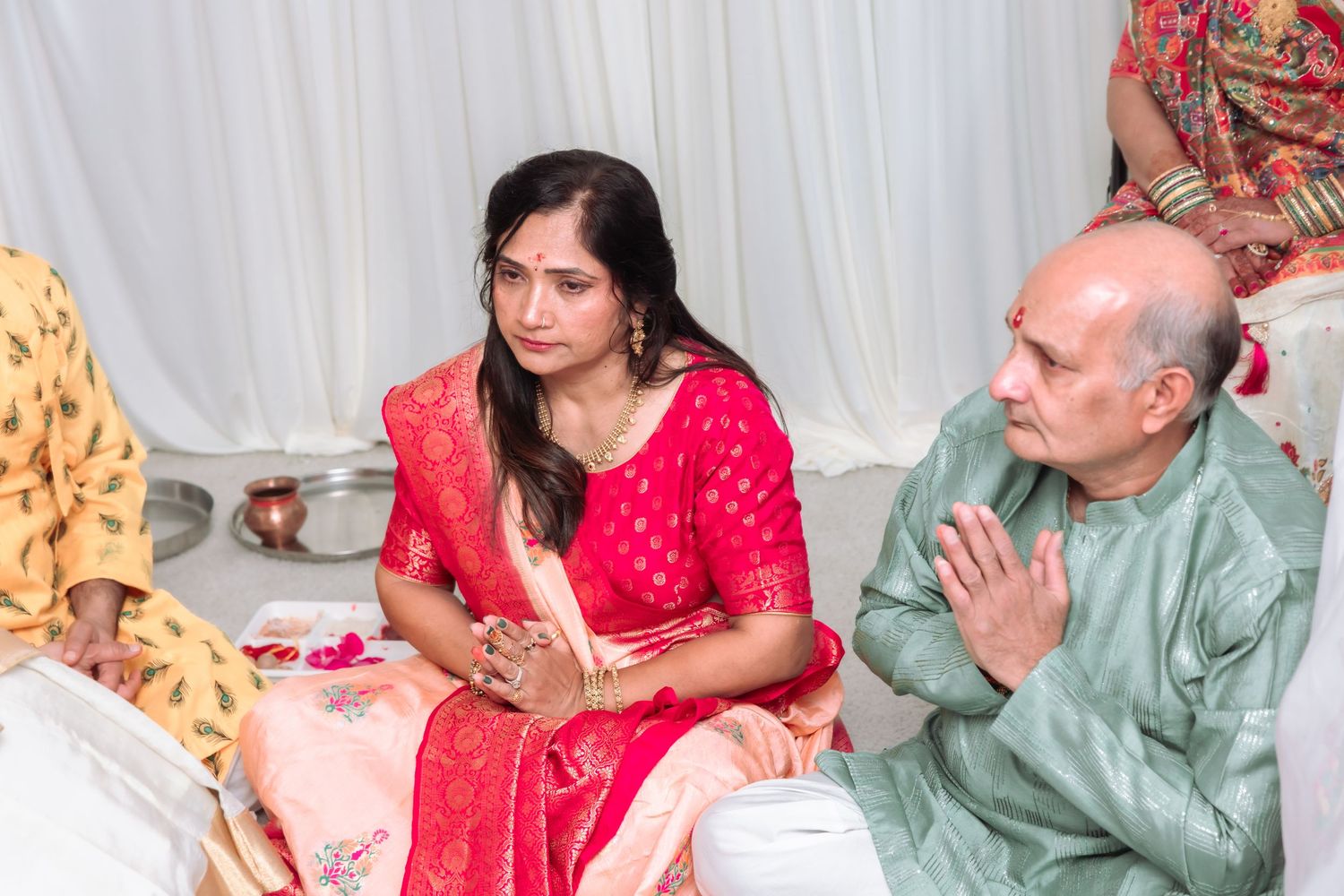 A woman in a pink salwar kameez and a bald man in sage green kurta sit during an Indian wedding ceremony.