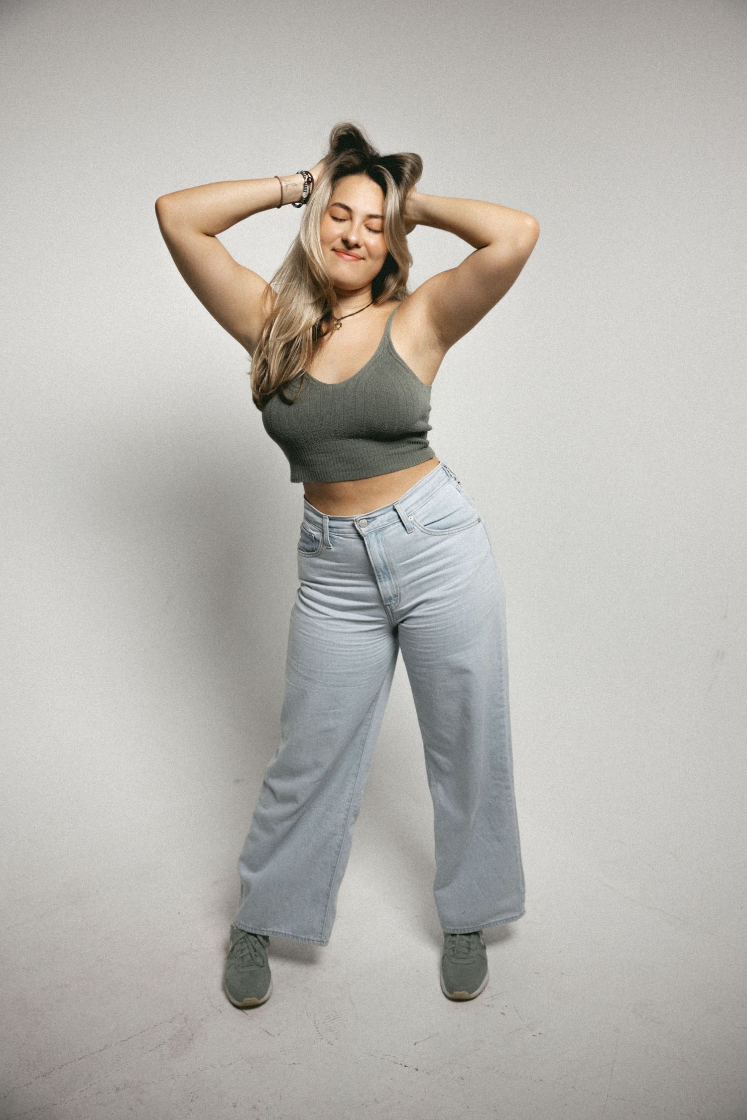 Young woman in gray crop top and wide-leg light blue jeans posing with hands in hair against white background.