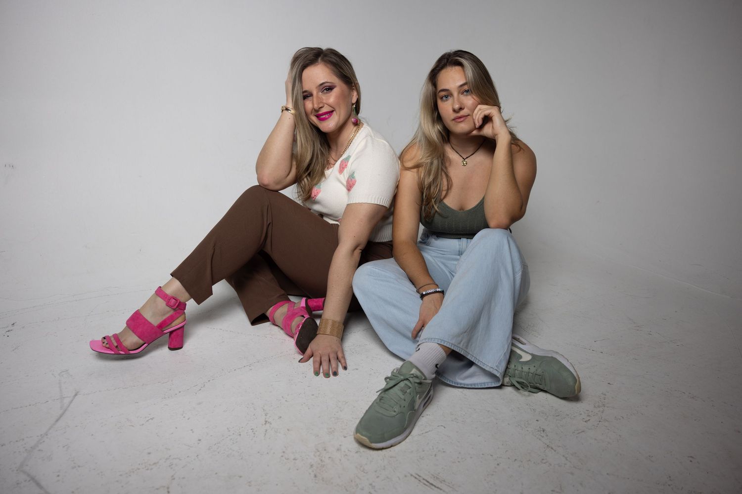 Two women sitting on a studio floor, one in brown pants and pink heels, the other in blue jeans.
