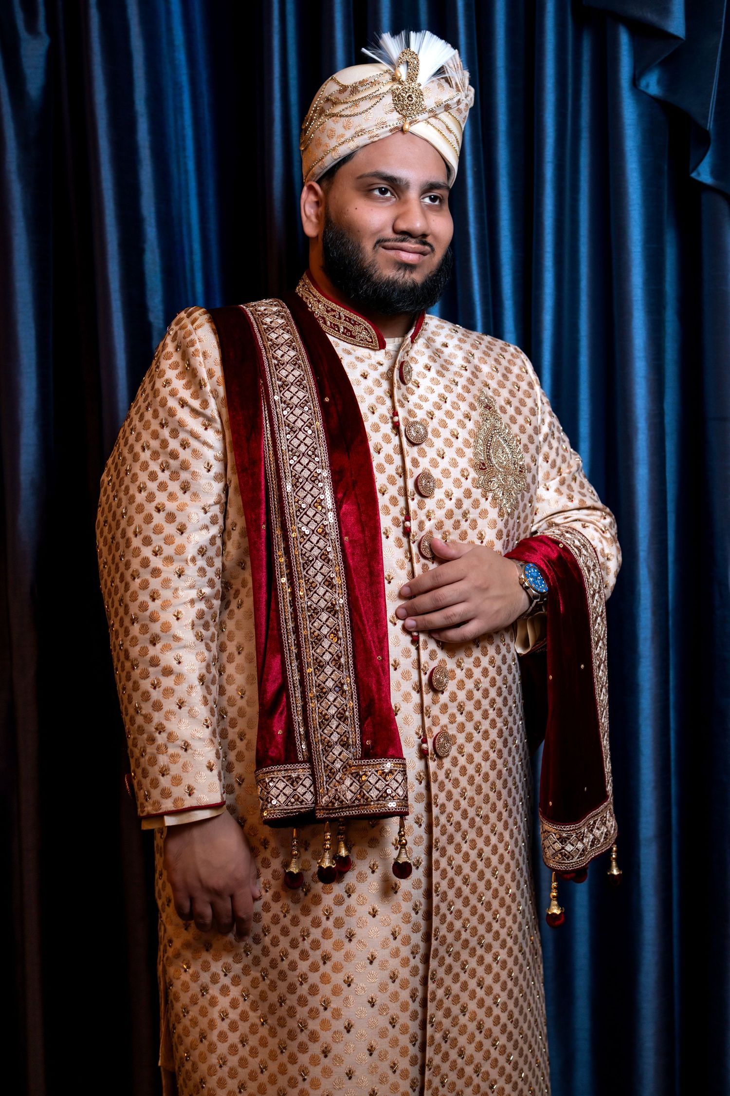 Indian groom in embroidered gold sherwani and turban posing with hand on chest before blue curtains.
