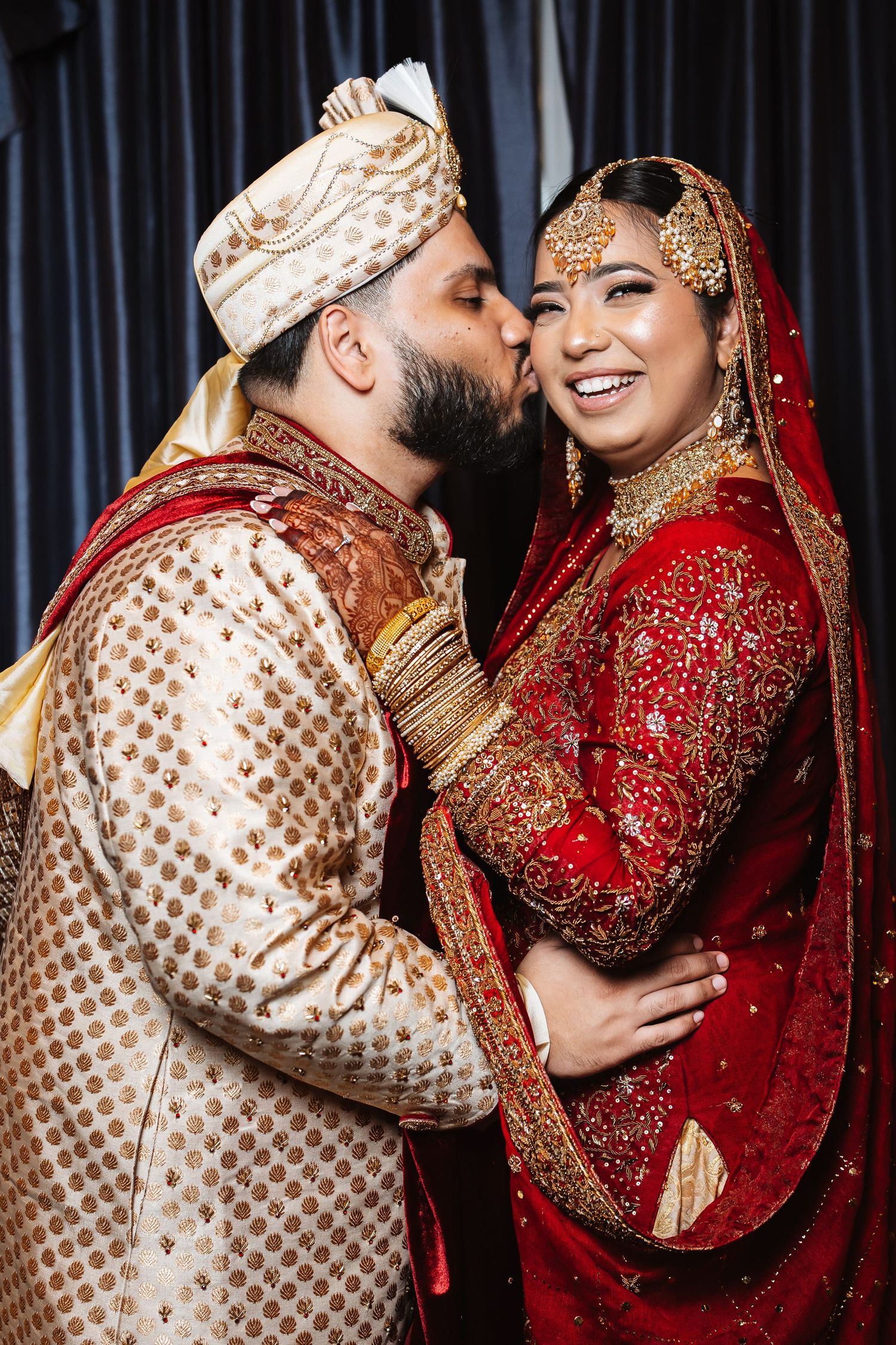 Indian groom in gold sherwani kissing smiling bride in red bridal lehenga on the cheek.