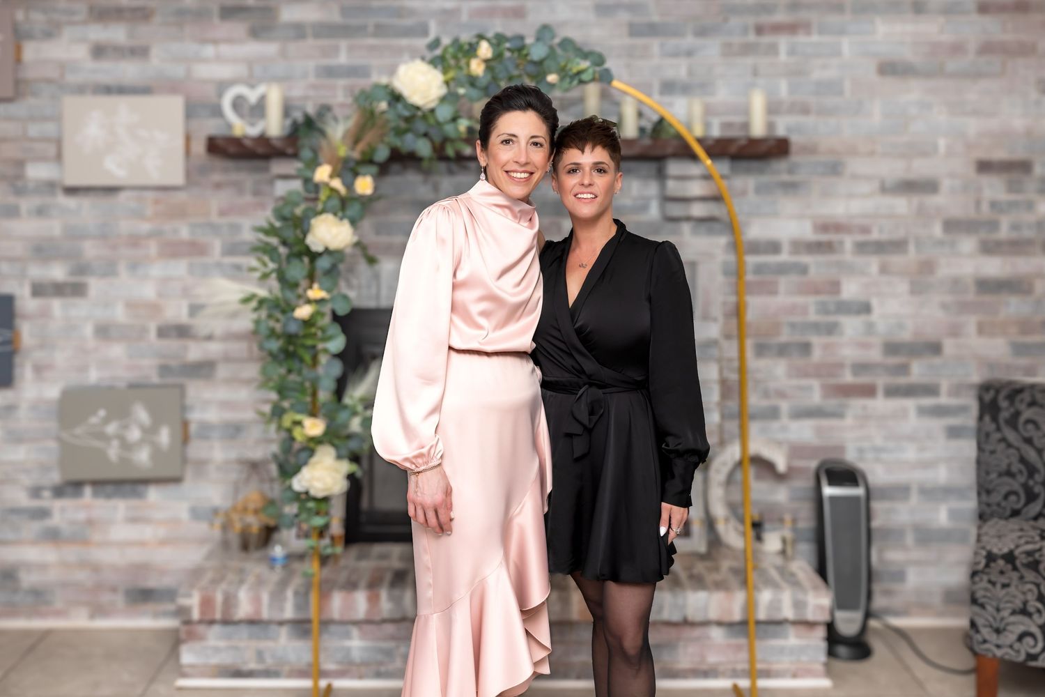Two women in elegant formal dresses smiling together in front of a gold floral arch backdrop.
