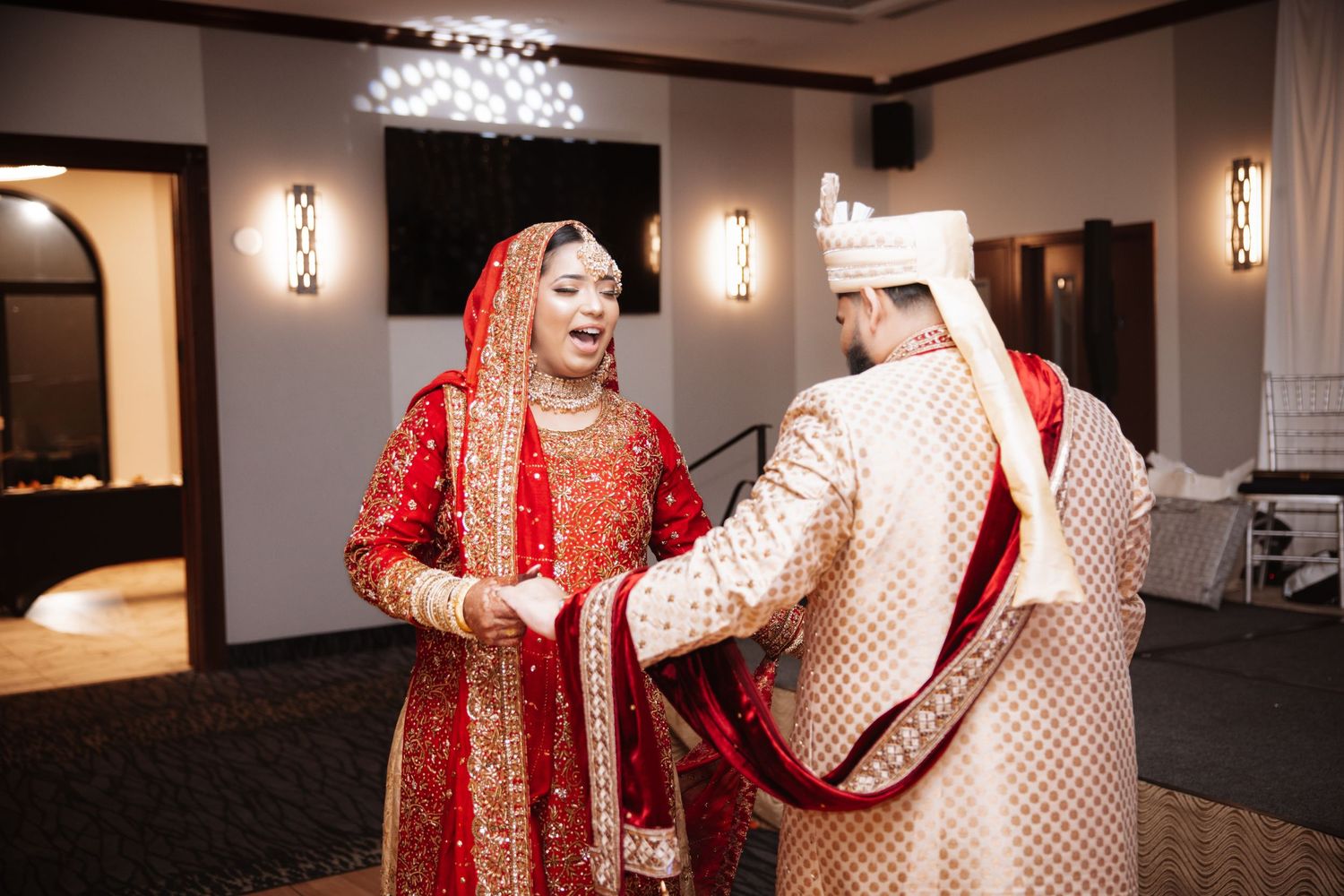 South Asian bride in red bridal lehenga laughing with groom in gold sherwani during ceremony.