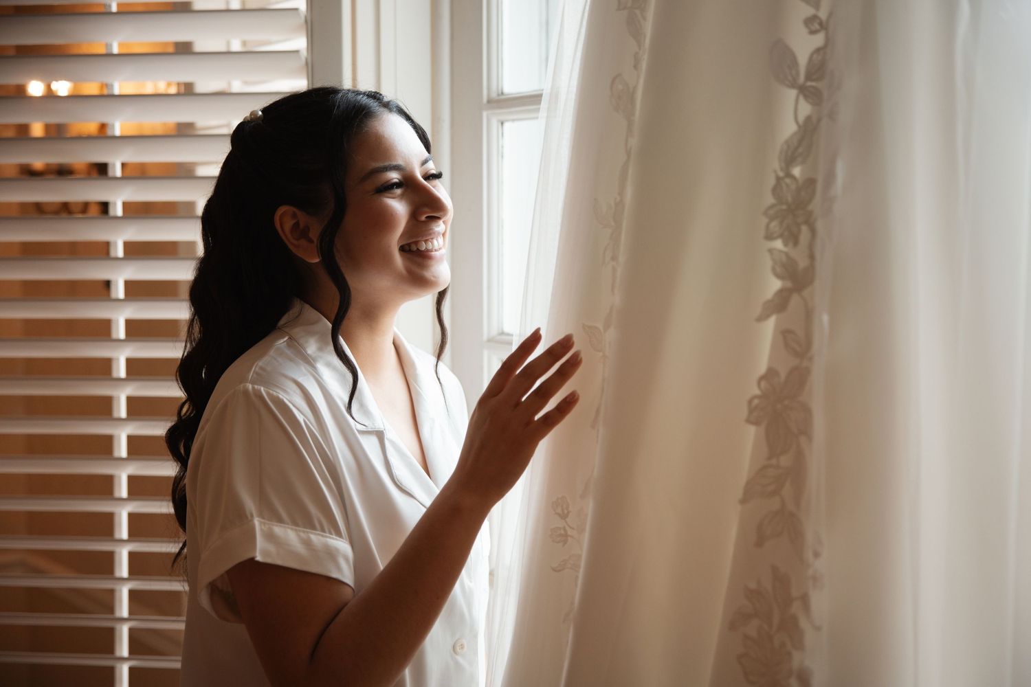 Happy dark-haired bride in white shirt smiling while touching her lace wedding dress hanging near a sunny window.