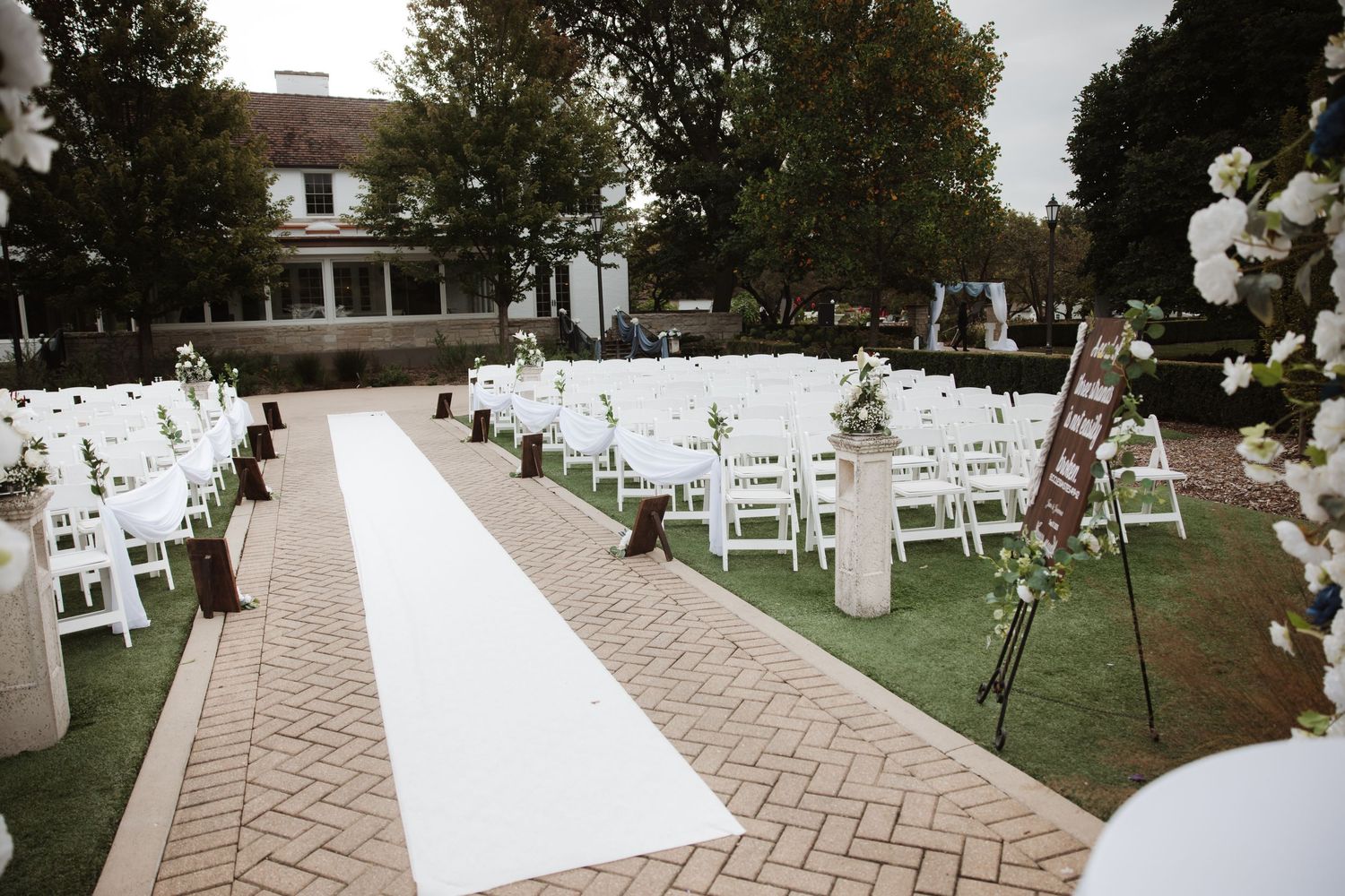 Outdoor wedding ceremony with white aisle runner, white chairs, floral arrangements, and wooden sign on brick path.