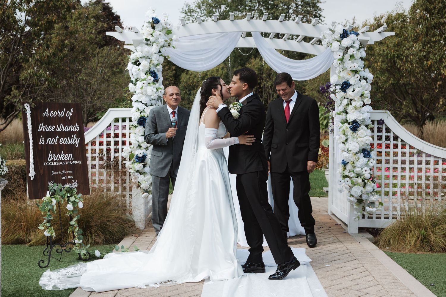 Bride and groom share first kiss under white floral arch at outdoor wedding ceremony.