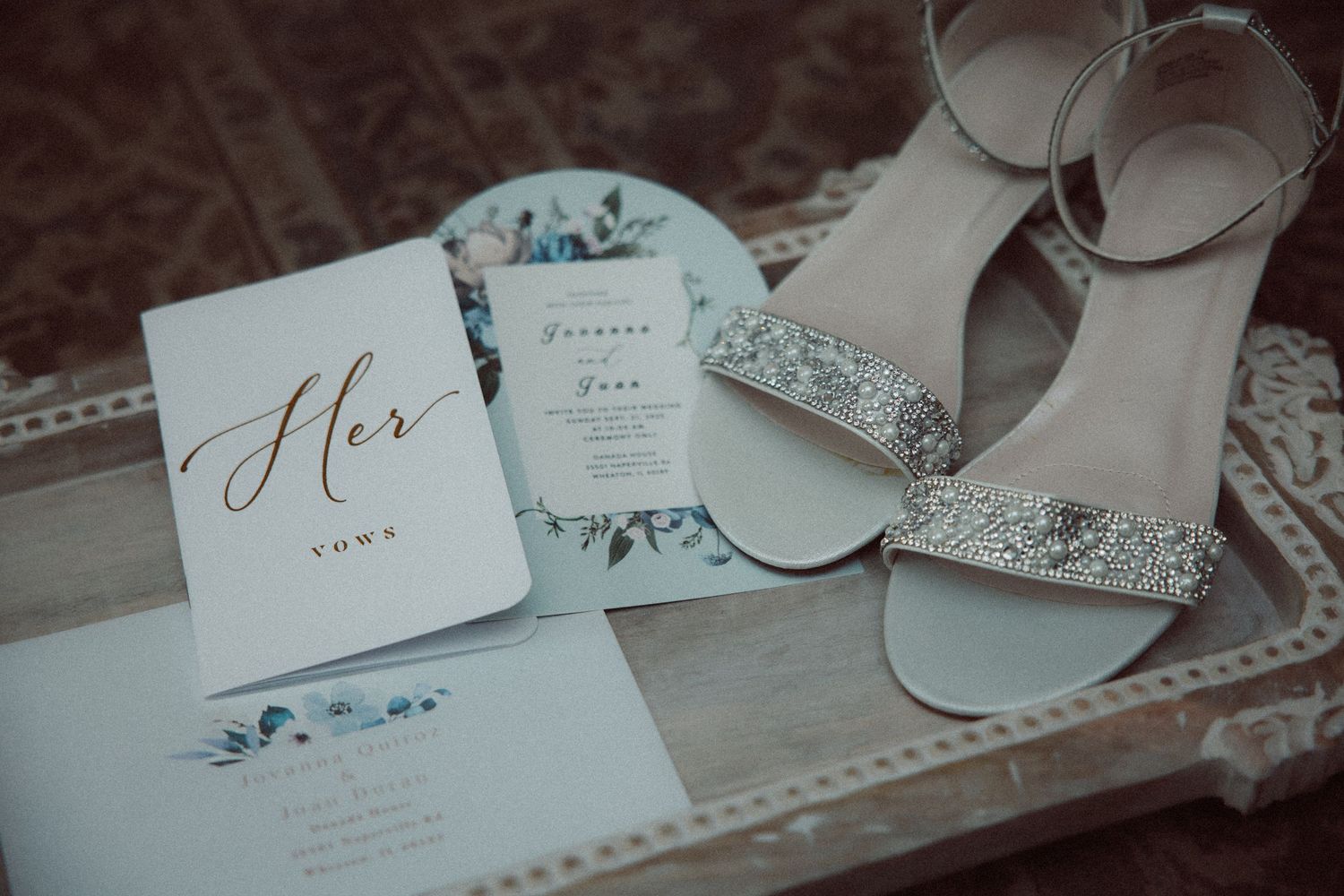 Elegant wedding detail shot of sparkly bridal sandals beside Her vows booklet and floral wedding invitation.
