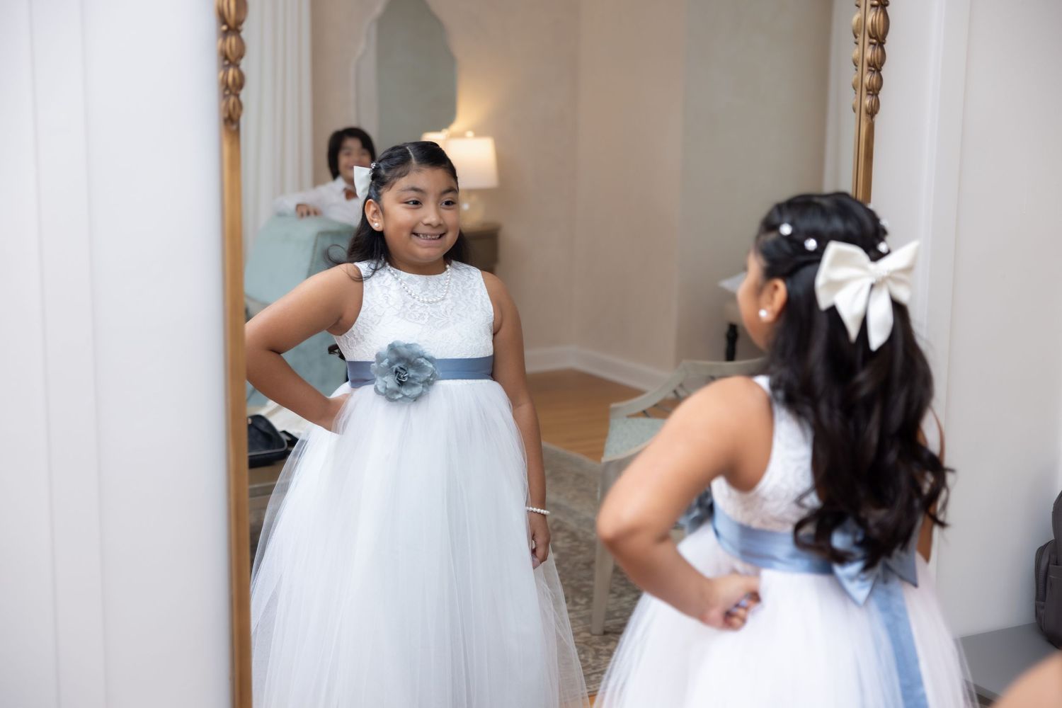 Young flower girl in white and blue dress admiring herself in a large ornate mirror.
