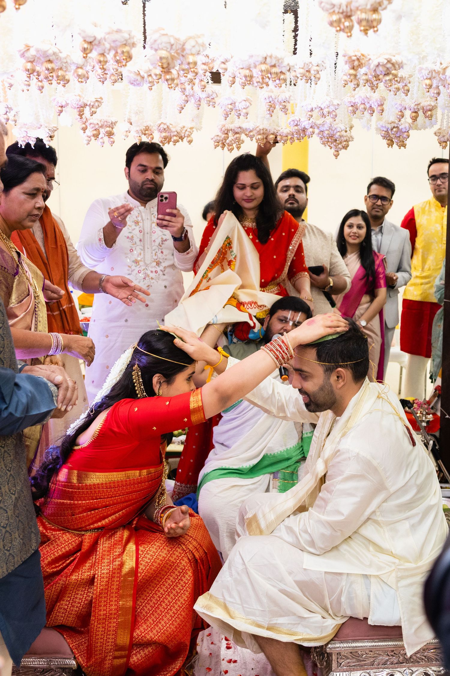 Bride in red saree applying tilak to groom during traditional Indian Bengali wedding ceremony with guests.
