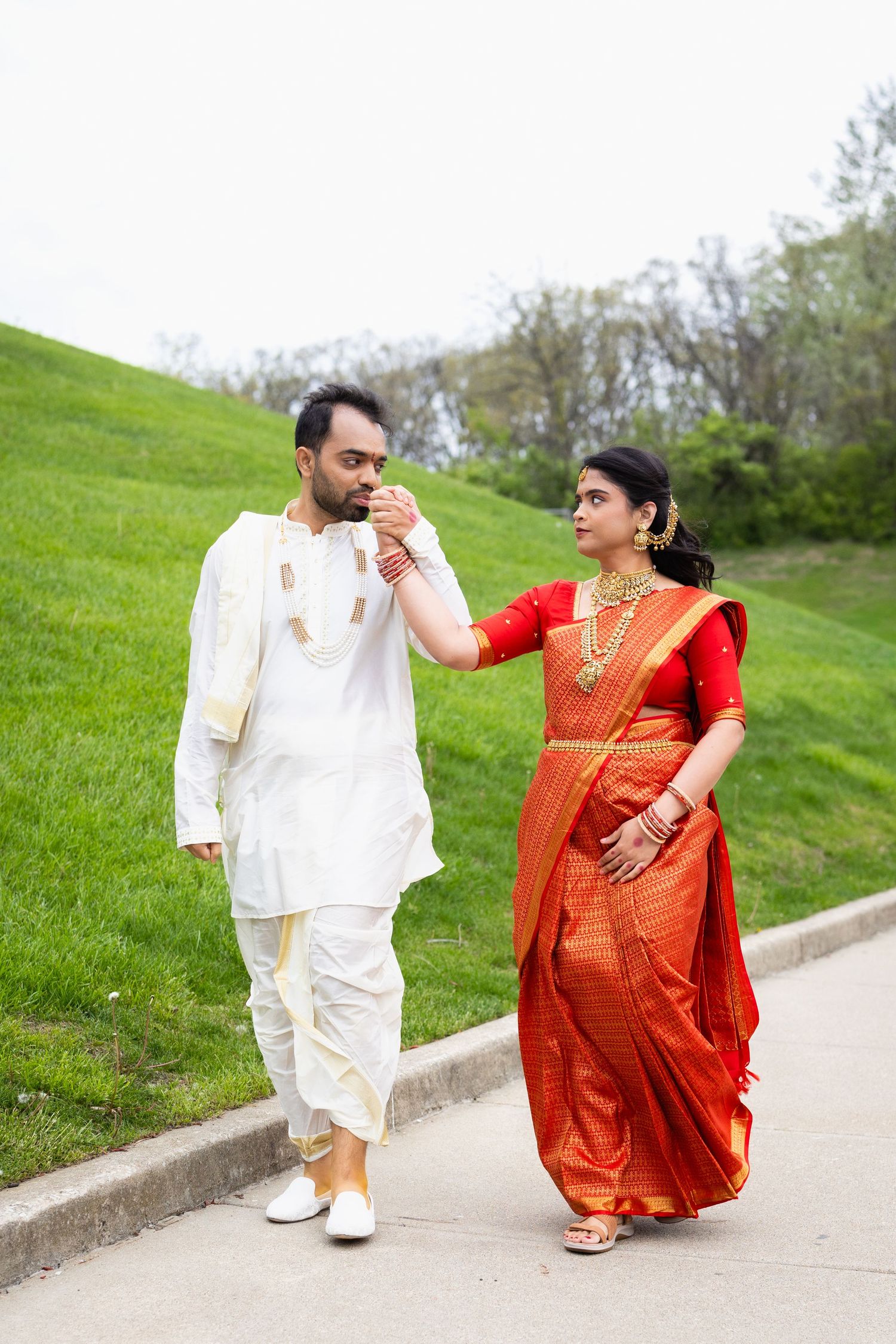 Indian groom in white kurta kissing bride's hand as she wears a red and gold saree outdoors.