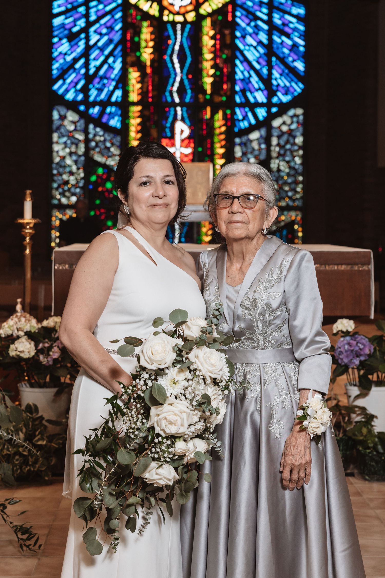 Bride in white gown holds white rose bouquet posing with elderly grandmother in silver dress at church altar.
