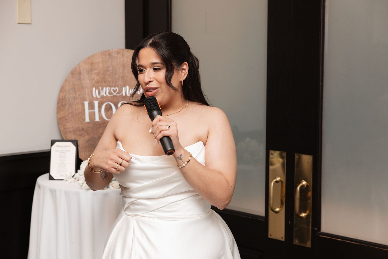 Bride in strapless white gown speaking into microphone at wedding reception.