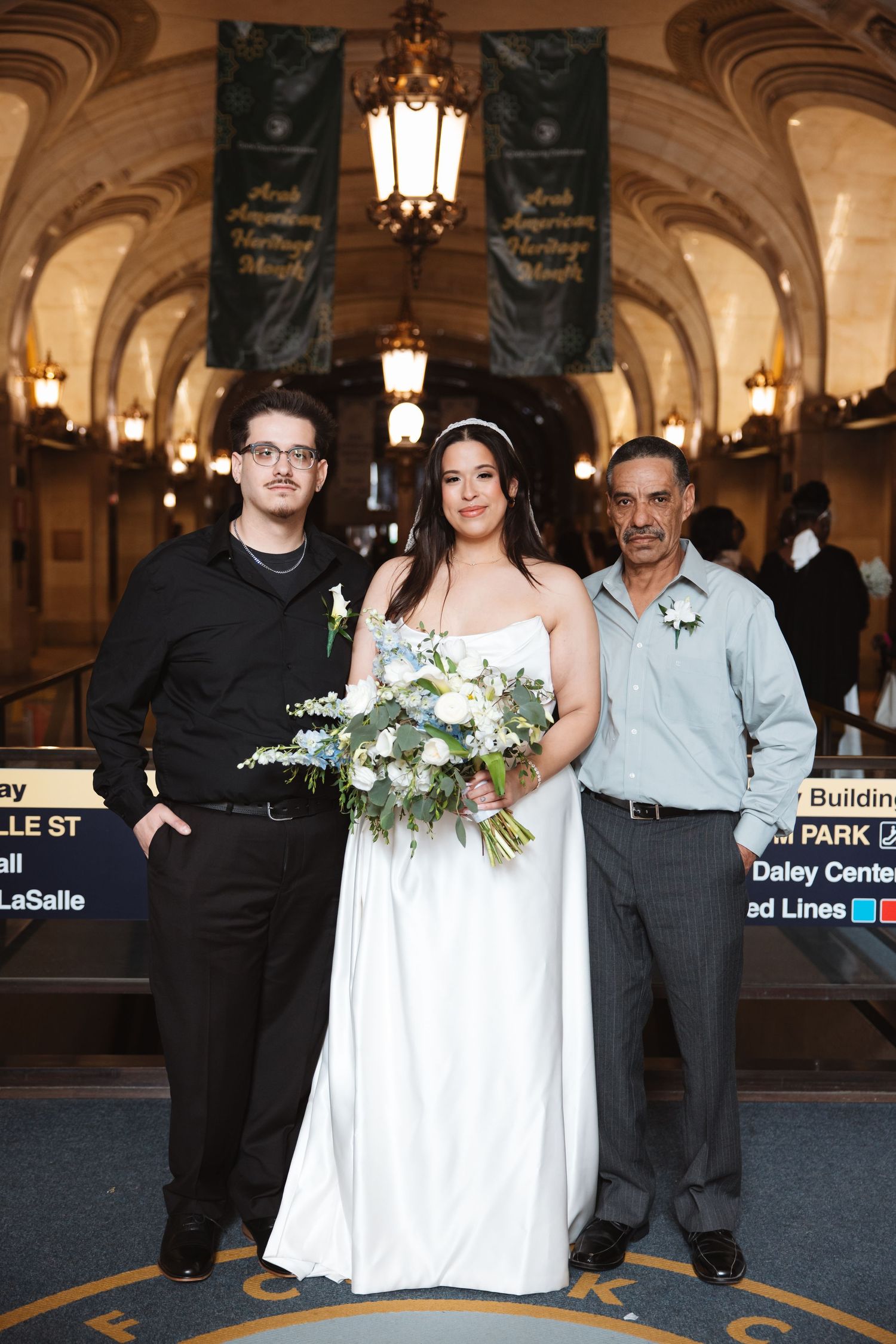 Bride in white gown holds bouquet flanked by two men in formal attire inside ornate building.
