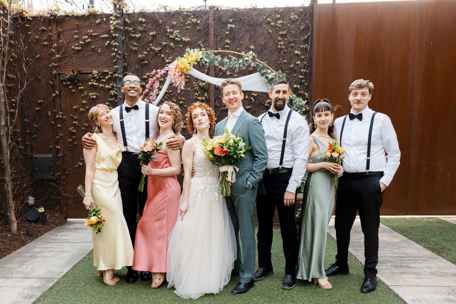 Wedding party poses formally in front of a floral arch, bridesmaids in pastel gowns and groomsmen in suspenders.