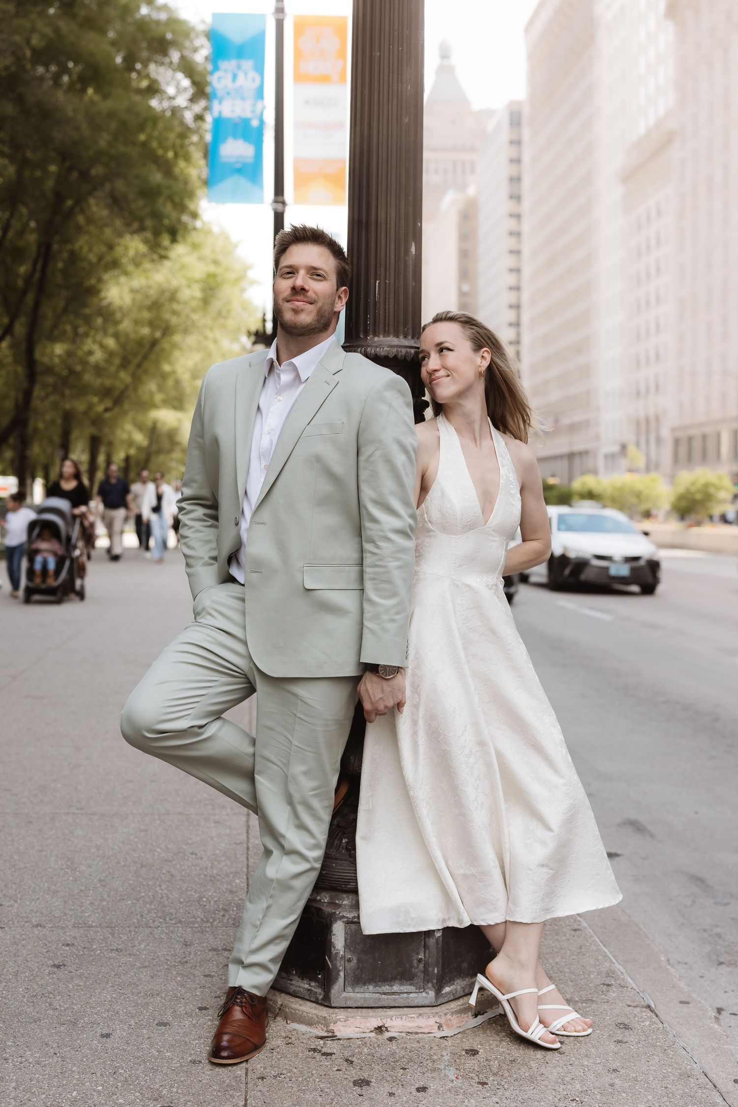 Wedding couple in sage suit and white halter dress pose against city street lamp post.