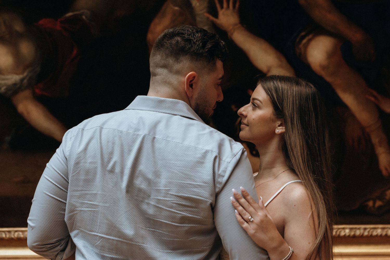 Couple sharing an intimate moment in front of a large classical painting in a museum gallery.