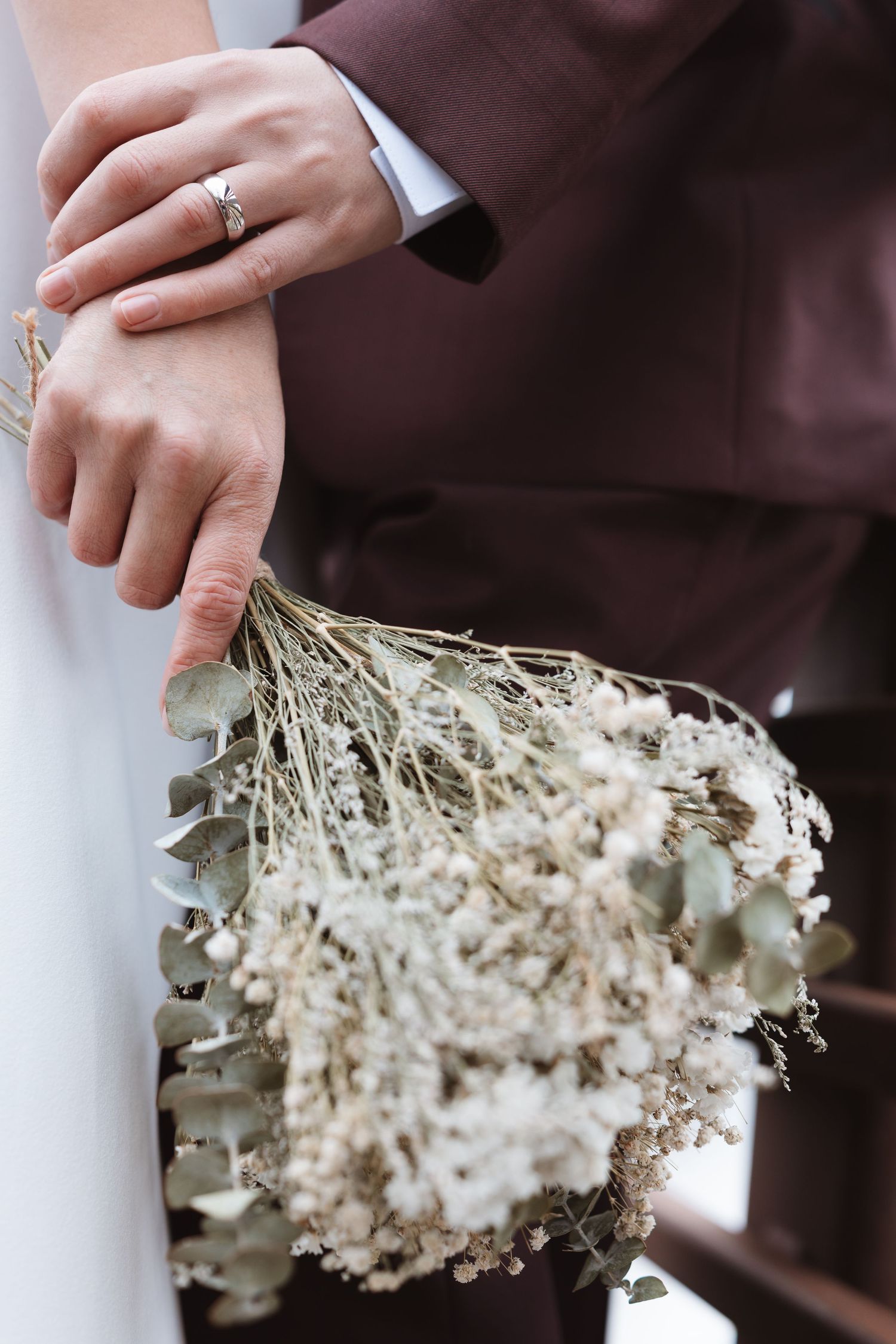 Bride and groom holding a rustic dried flower bouquet with eucalyptus, showcasing an elegant ring on the bride's finger.