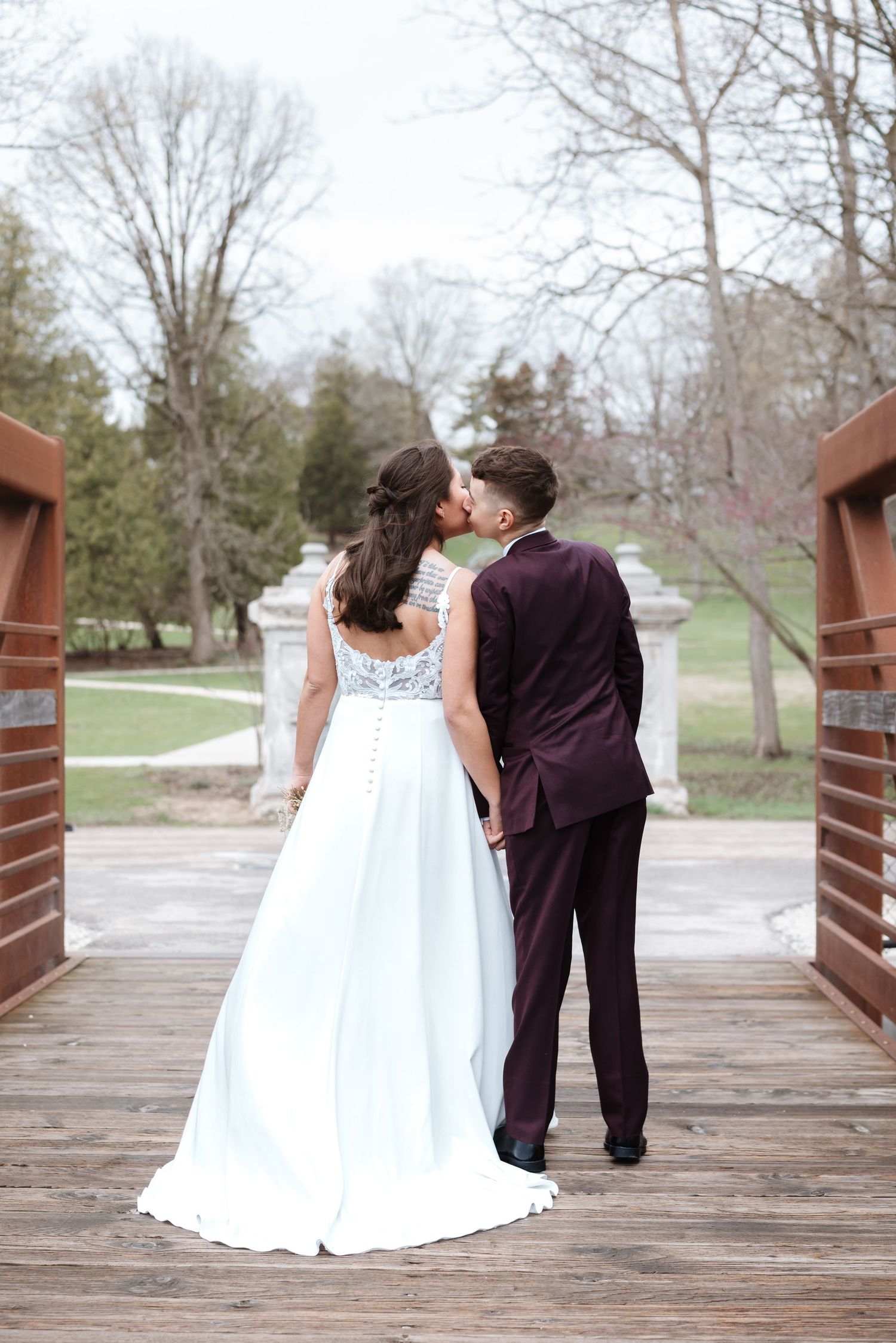 Bride and groom kissing on a wooden bridge in a park, bride in white gown, groom in burgundy suit.