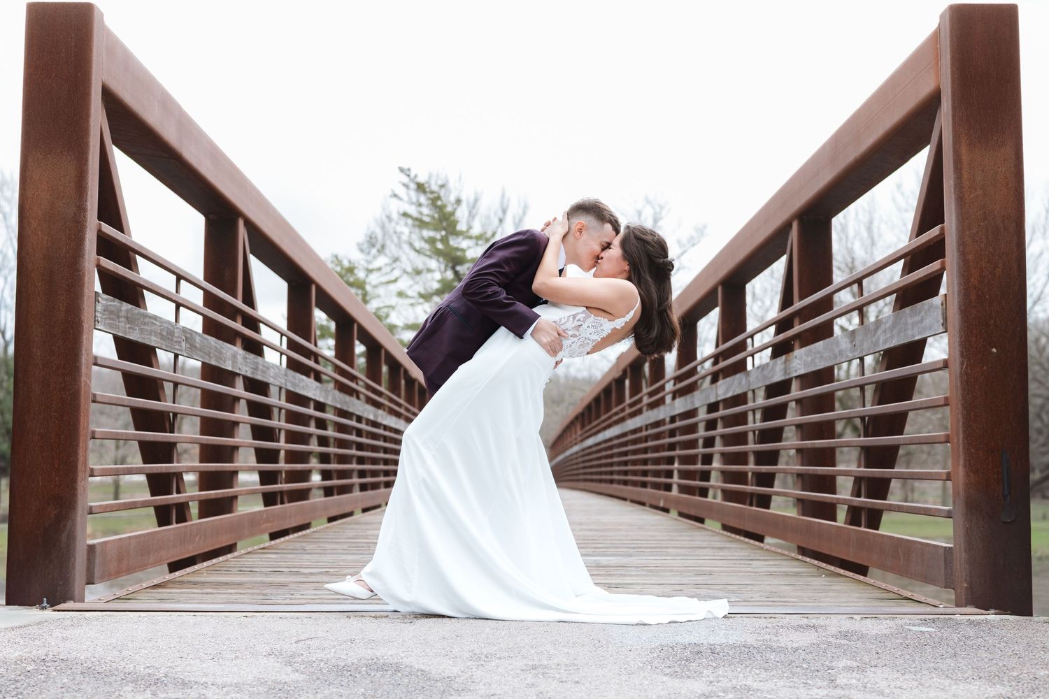 Bride and groom share a romantic dip kiss on a wooden bridge surrounded by trees.