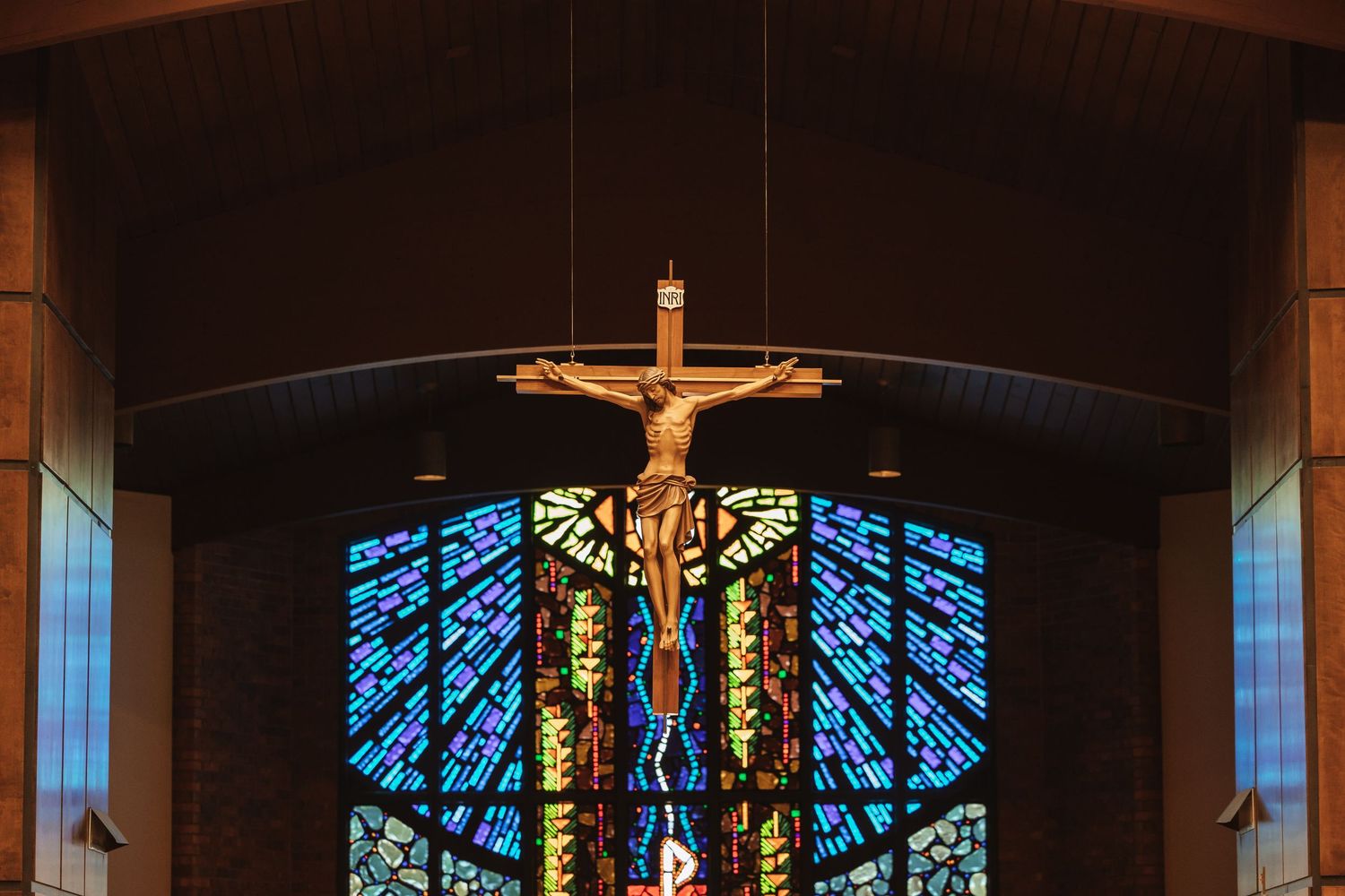 Crucifix hanging above colorful stained glass windows inside a Catholic church sanctuary.