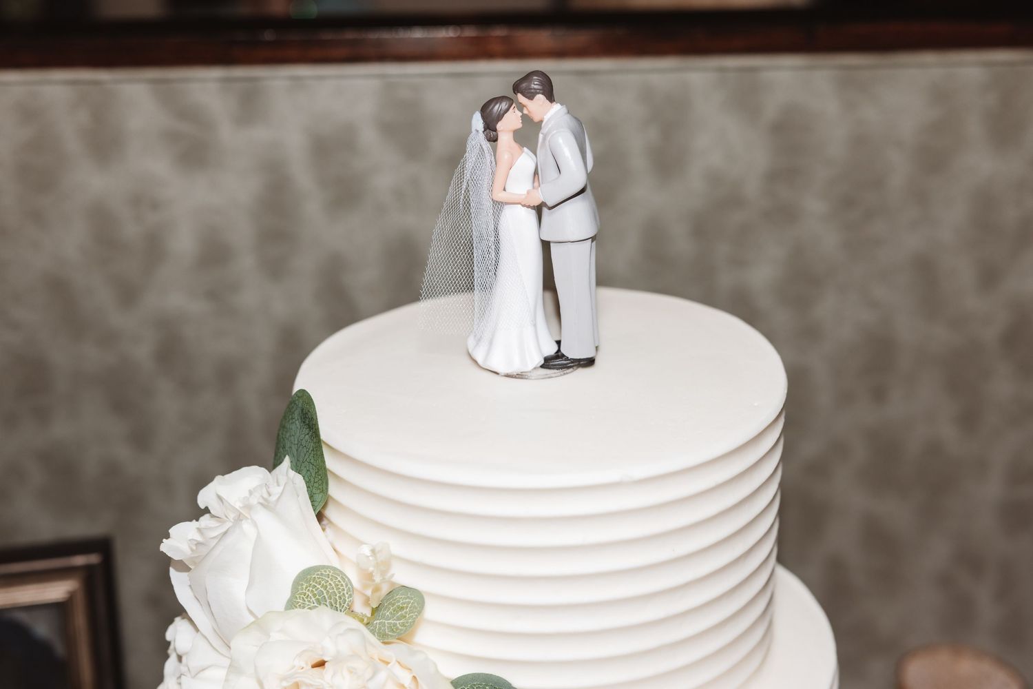 White tiered wedding cake with bride and groom cake topper and white floral decorations.