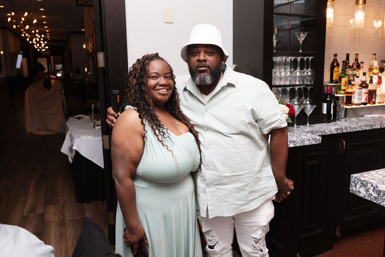 Couple smiles together at wedding reception, woman in sage green dress and man in white outfit.