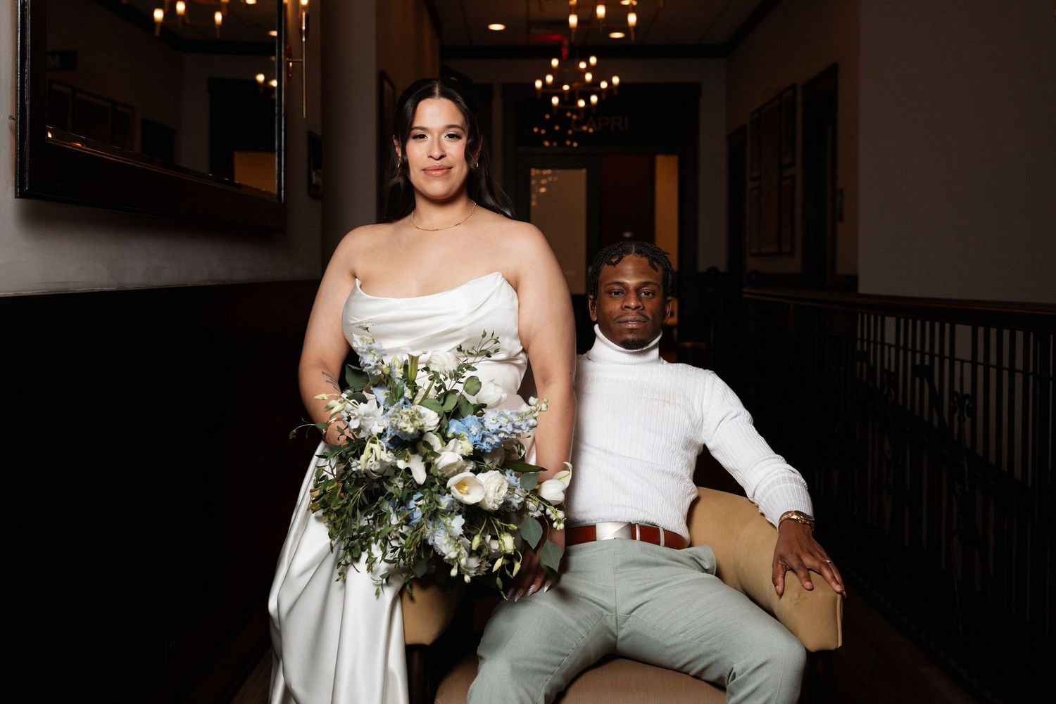 Bride holding blue and white bouquet standing beside groom seated in armchair.