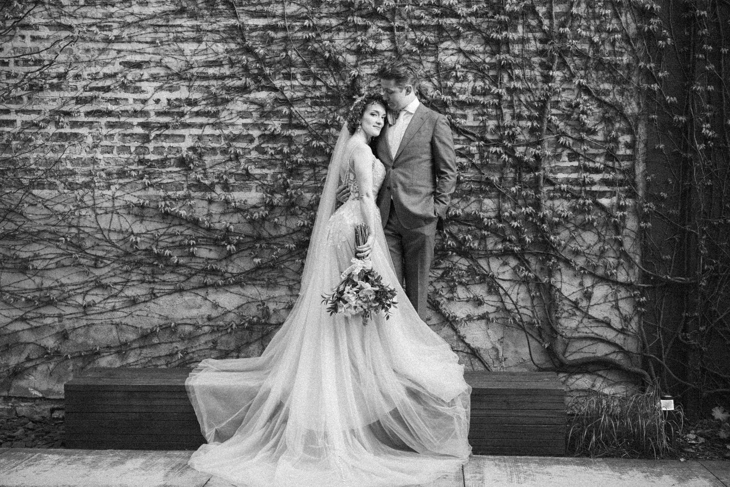 Bride and groom pose romantically in black and white photo against ivy-covered brick wall with flowing veil.