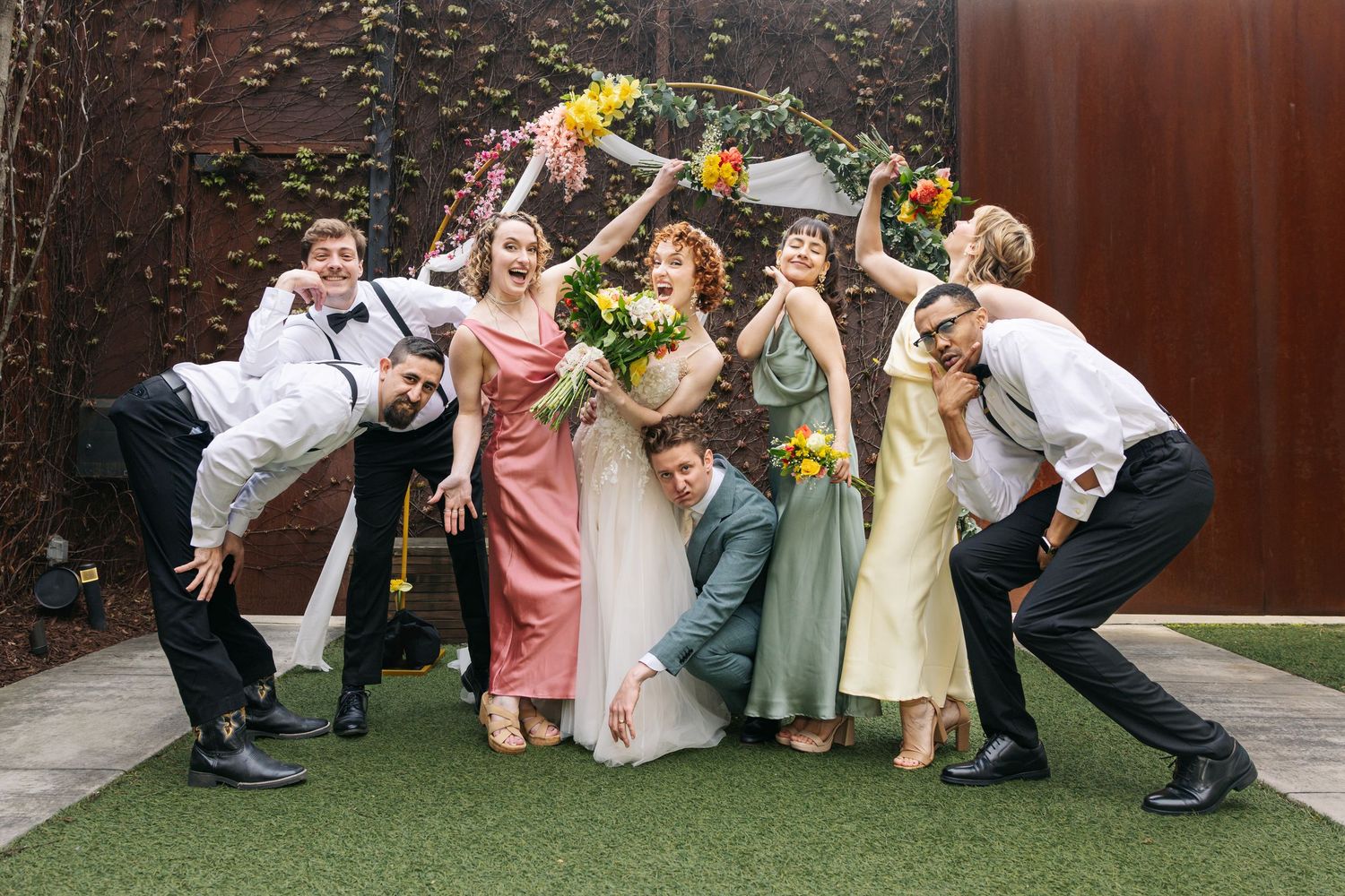 Fun wedding party photo with bride, groom and bridal party posing playfully with bouquets outdoors.