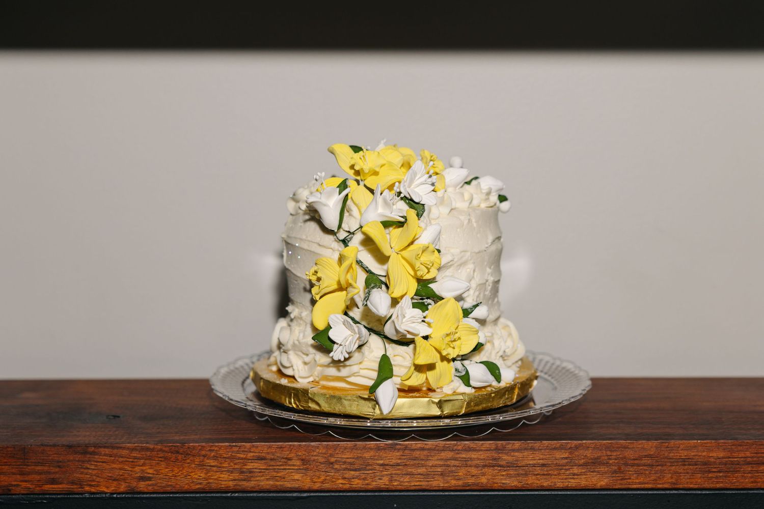 Two-tier white wedding cake decorated with yellow and white sugar flowers on wooden table.