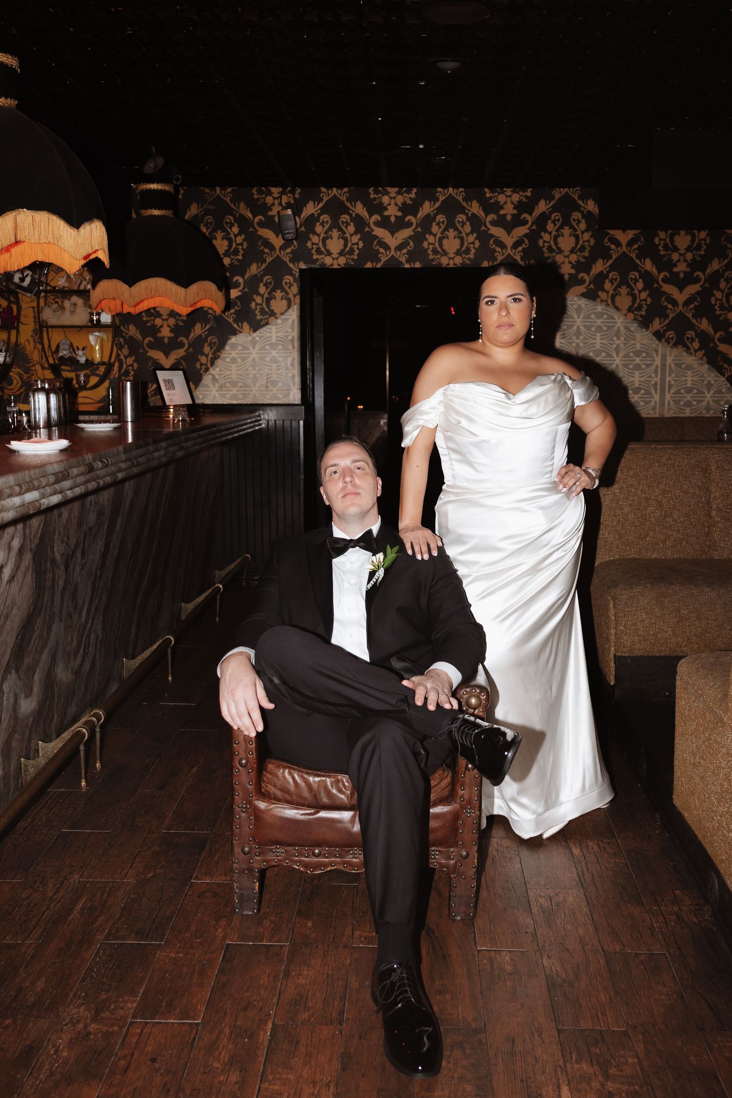 Bride and groom pose dramatically in a dark vintage bar setting, groom seated in leather chair.