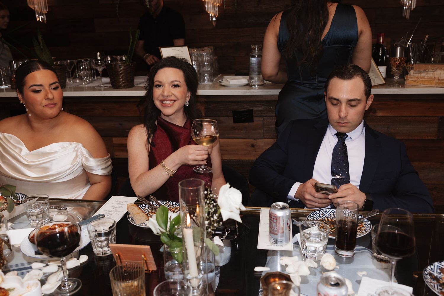 Wedding guests sit at dinner table, woman in white dress toasts with wine glass.
