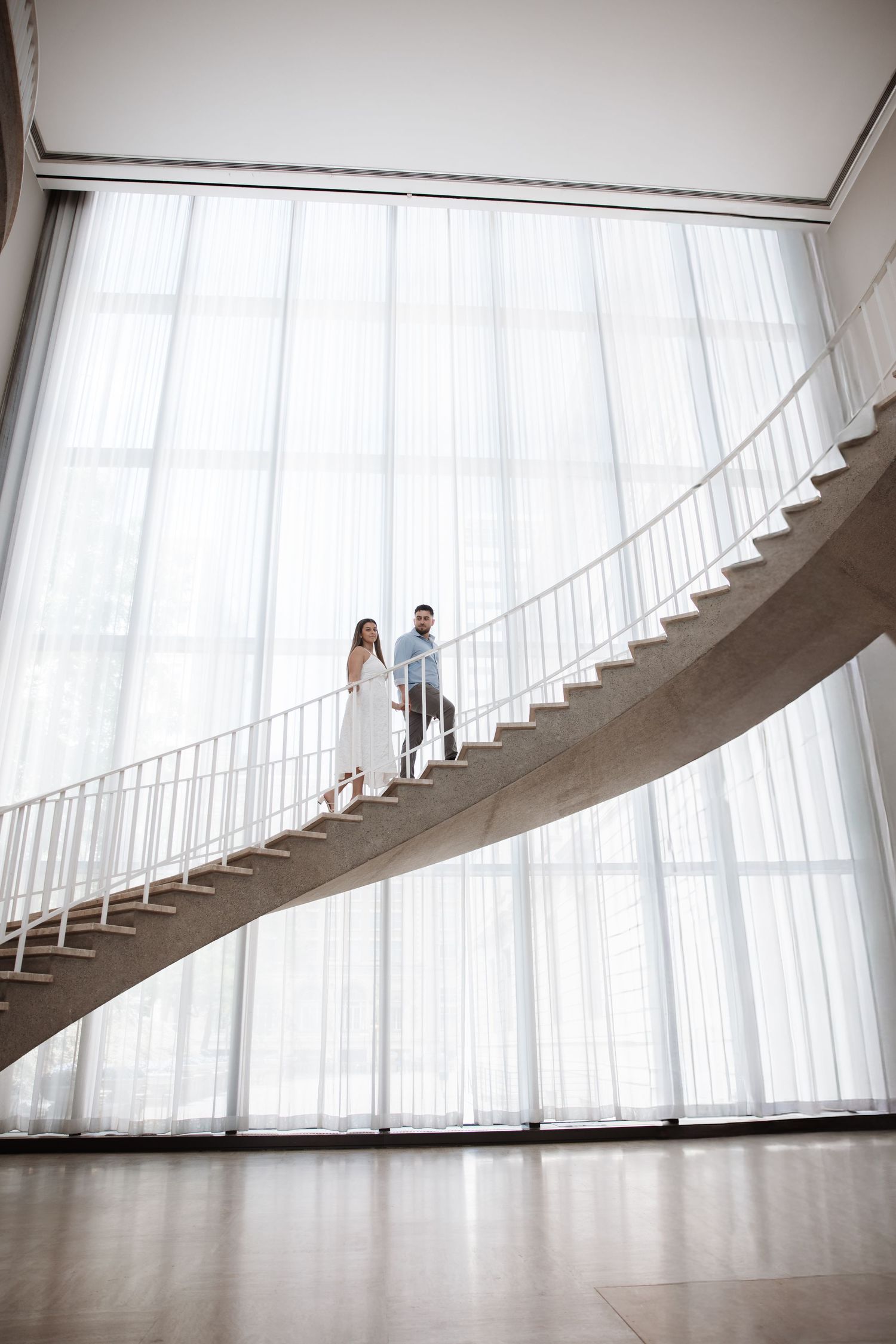 Couple standing together on a curved spiral staircase in front of large white curtained windows in modern building.