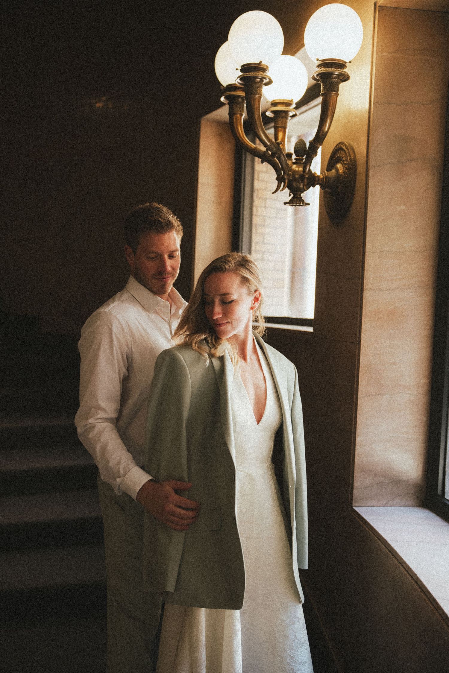 Groom tenderly drapes his suit jacket over bride's shoulders near elegant brass wall sconce.